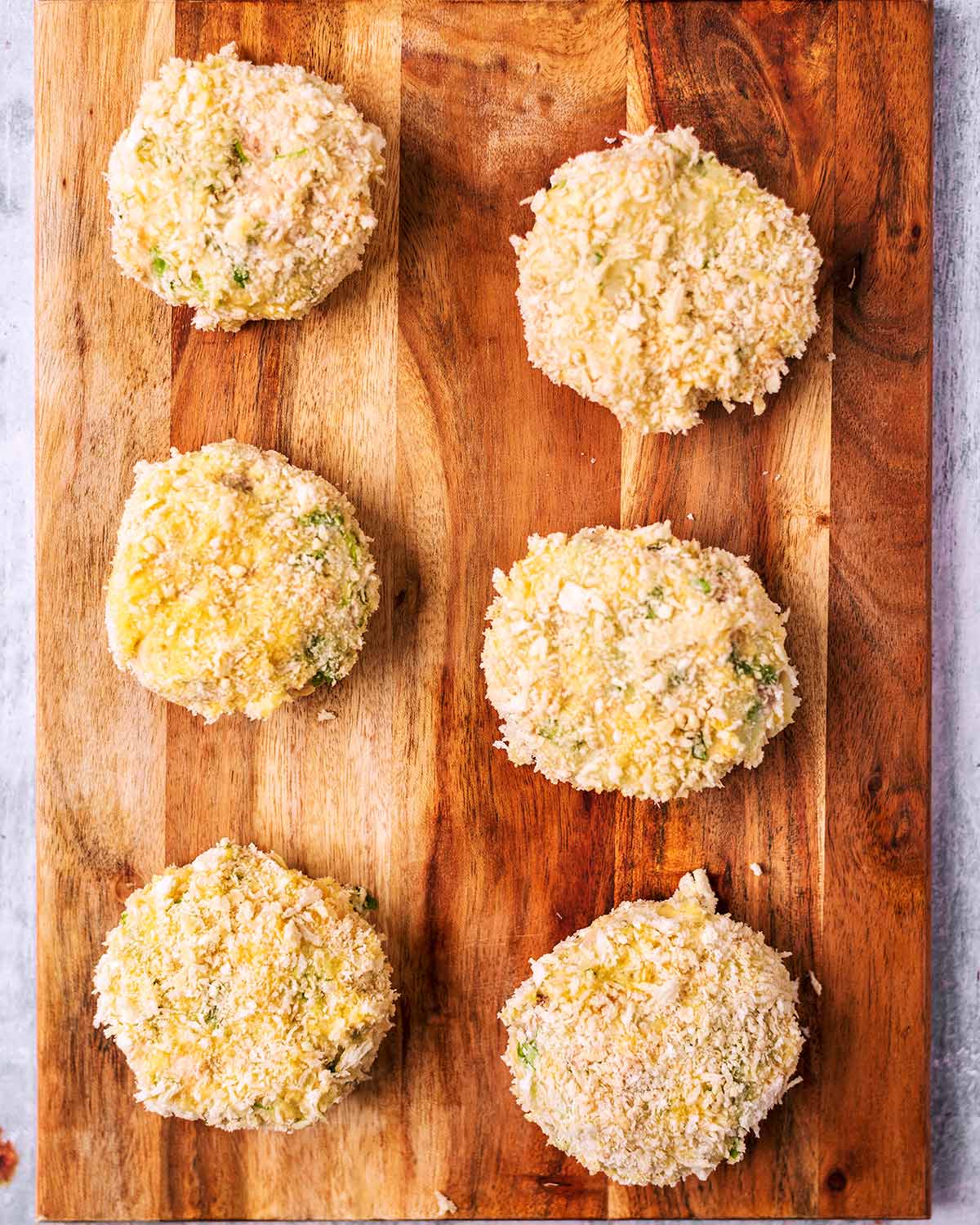 Six uncooked breaded fishcakes on a wooden board.