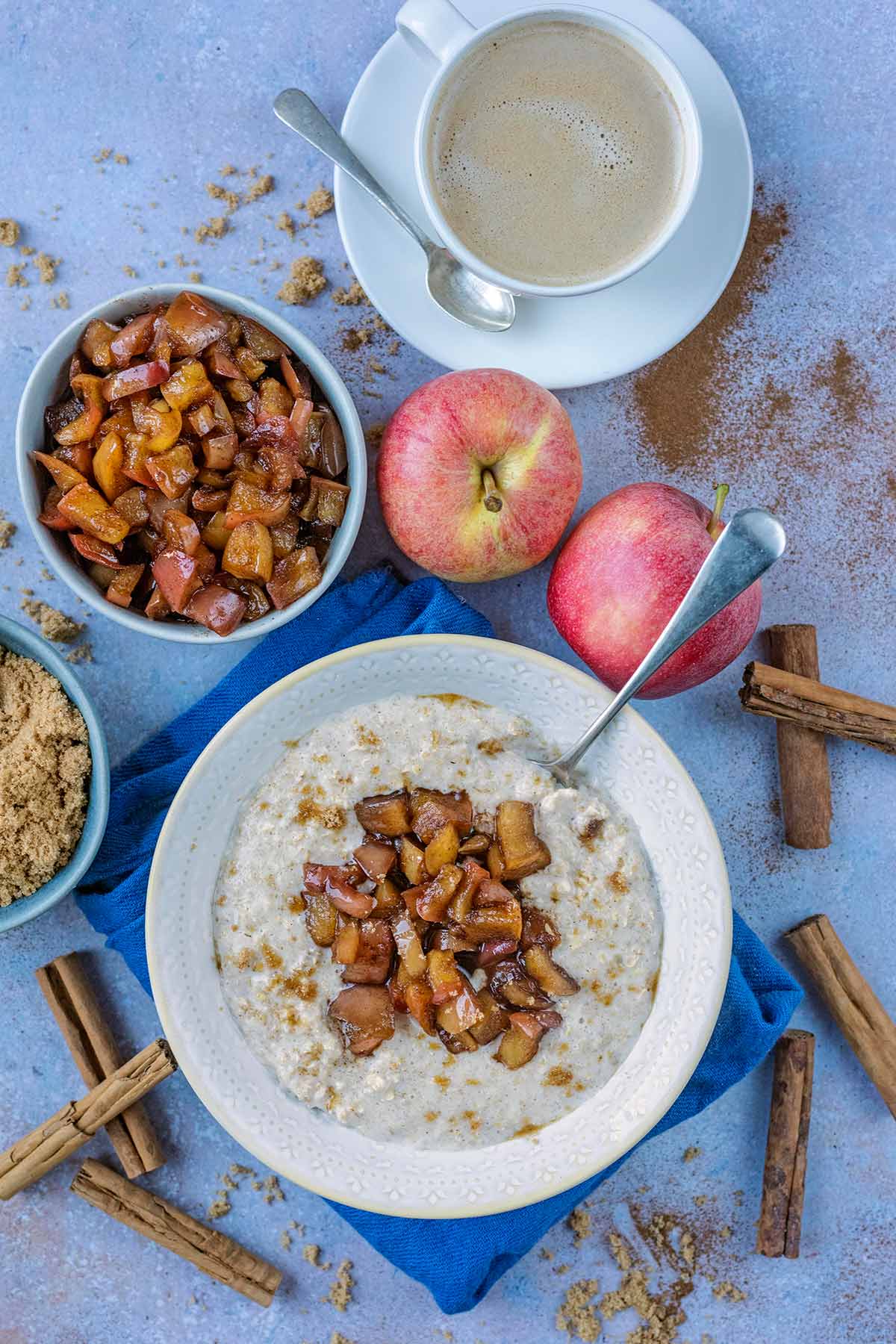 A bowl of porridge topped with cooked apples next to some apples and a cup of coffee.