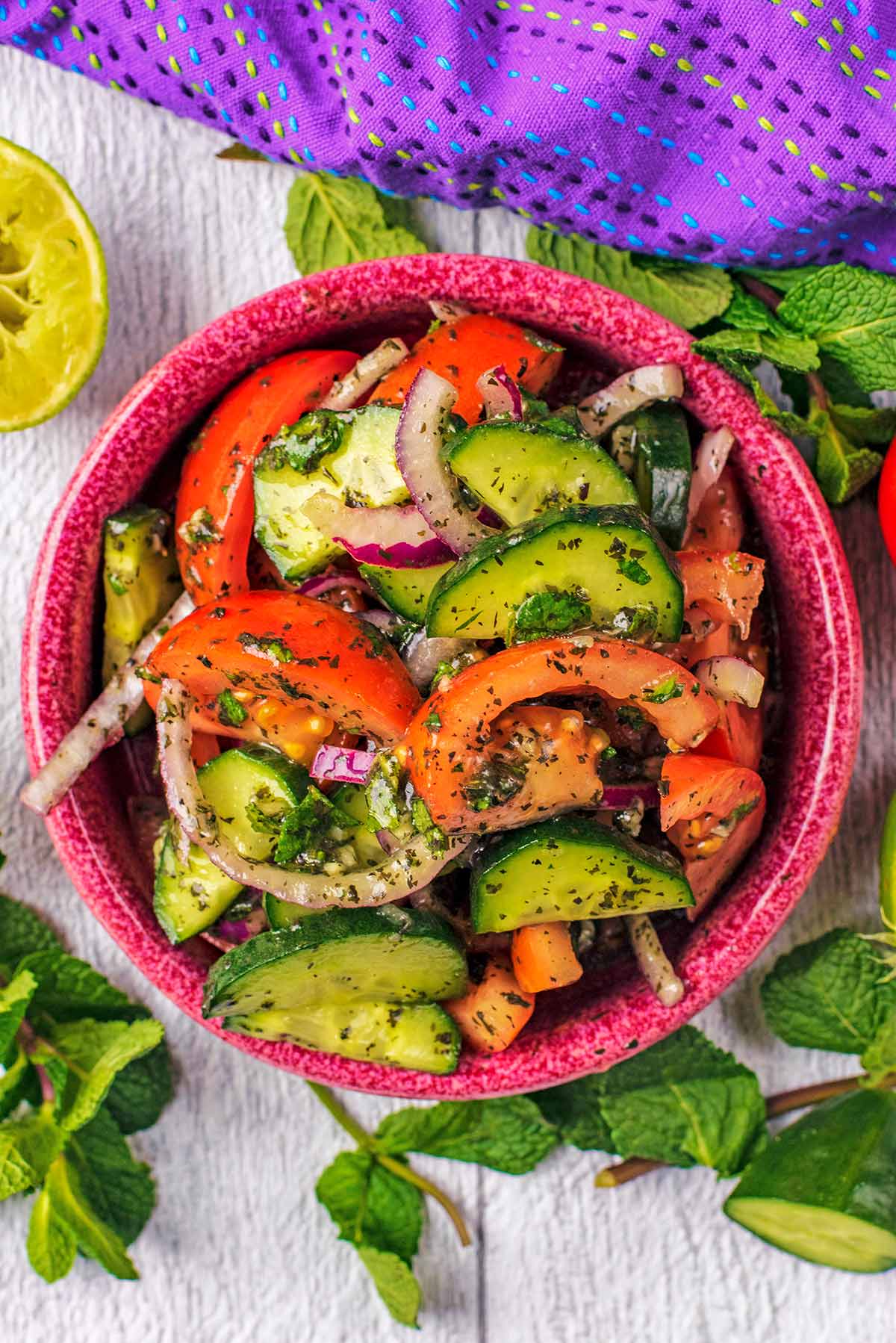 A bowl of sliced cucumbers, tomatoes and onions in a dressing, next to a purple towel on a wooden surface.