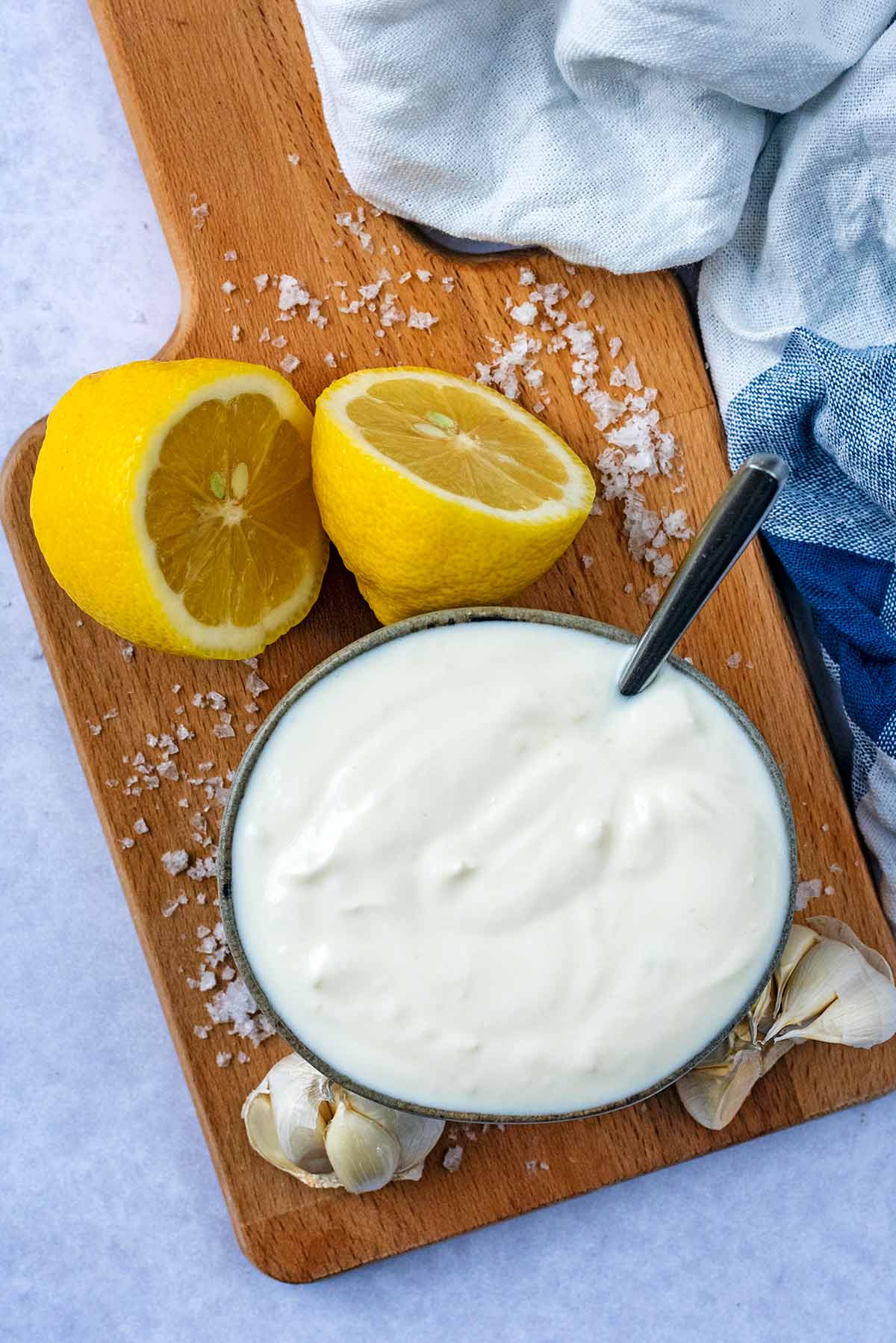 A bowl of yogurt sauce on a wooden board with garlic cloves and lemon halves.