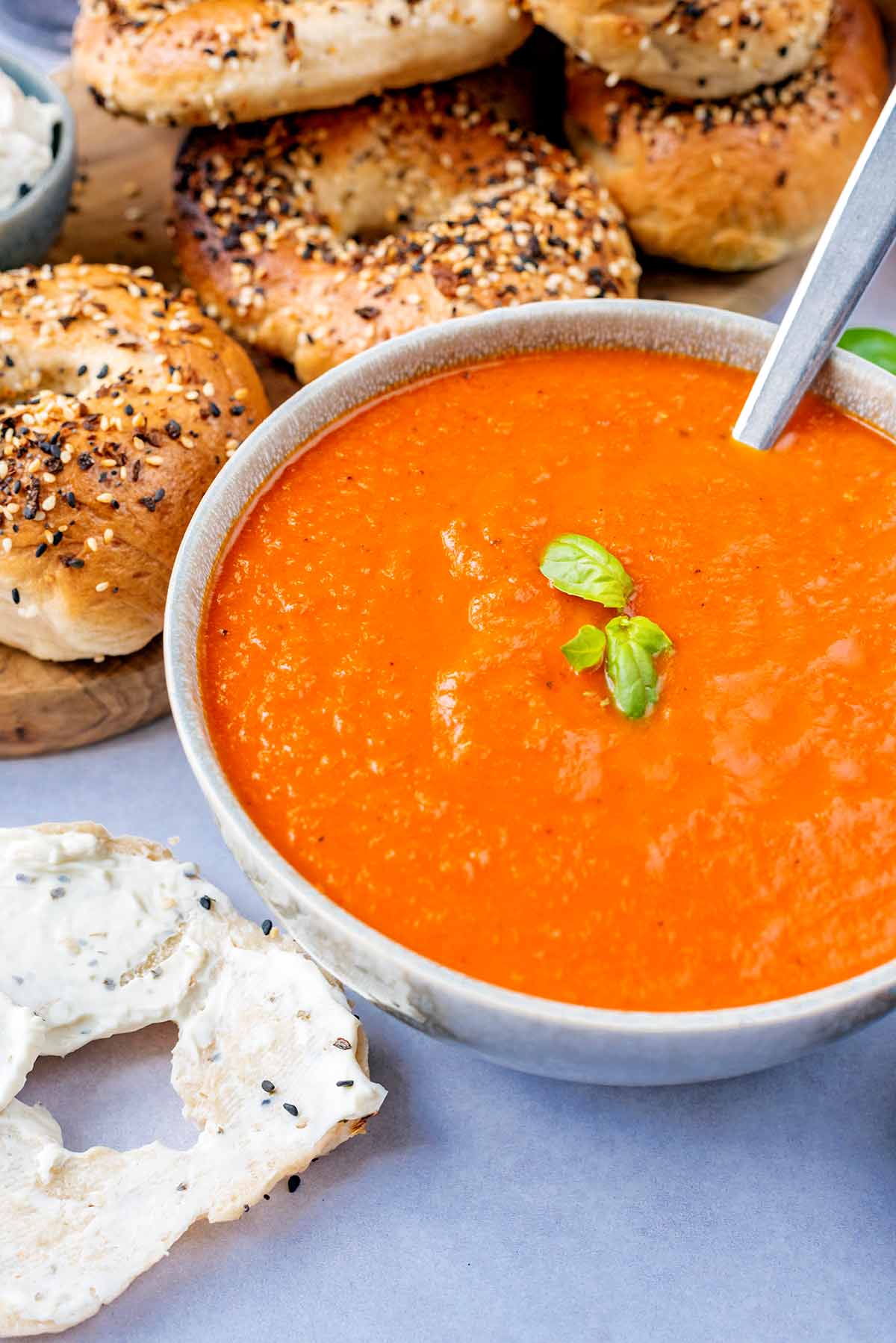 Tomato soup in a bowl with a spoon in front of some seeded bagels.