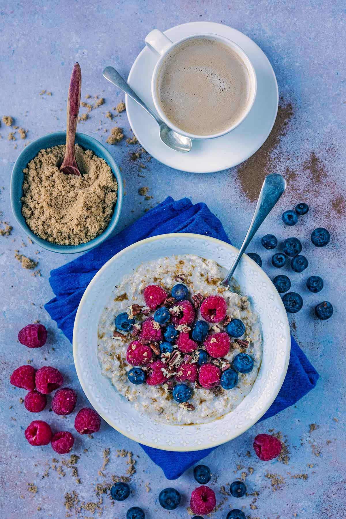 A bowl of porridge topped with berries, next to a bowl of sugar and a cup of coffee.