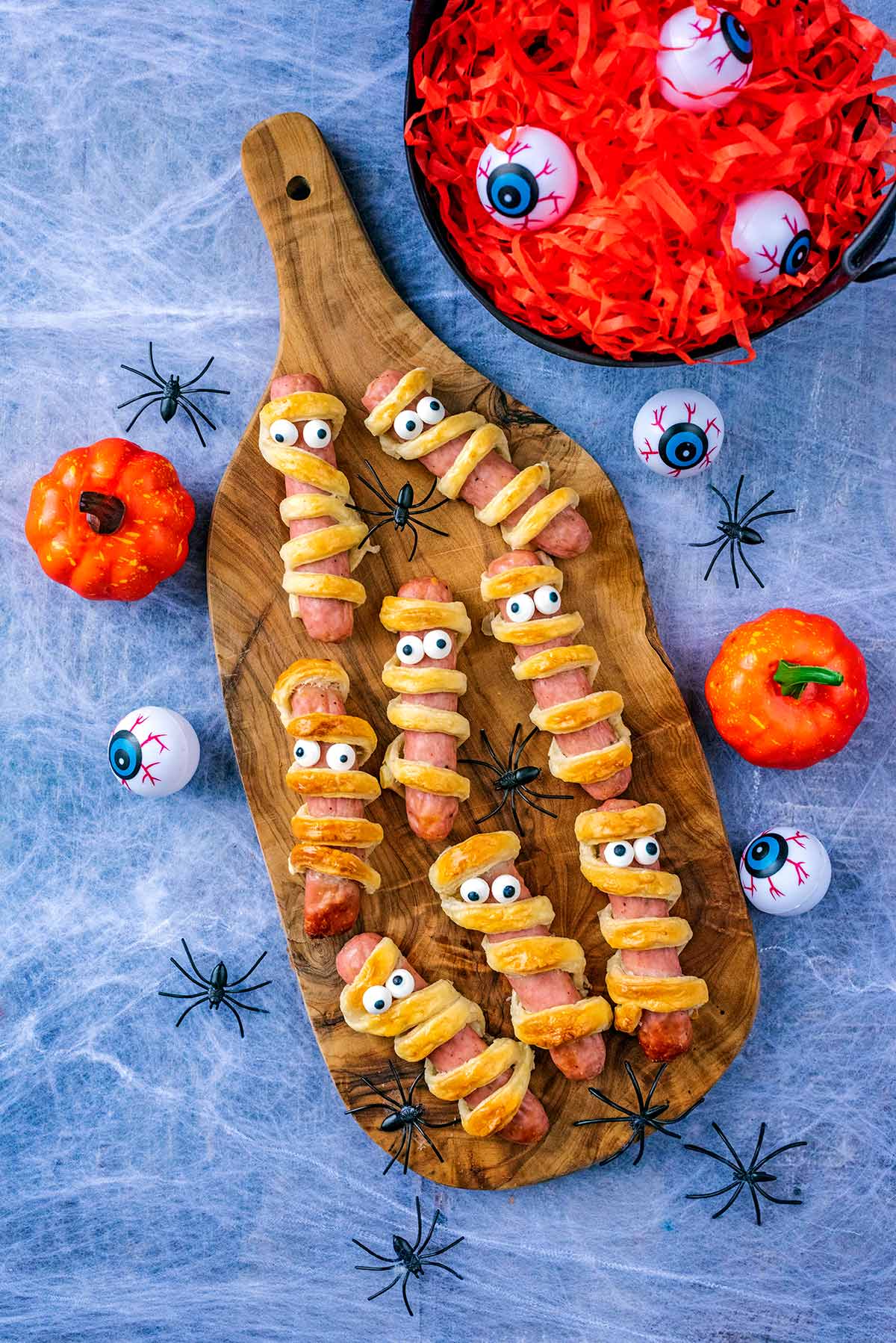 A wooden serving board with pastry wrapped sausages on it. Pumpkins, spiders and fake eyeballs surround it.