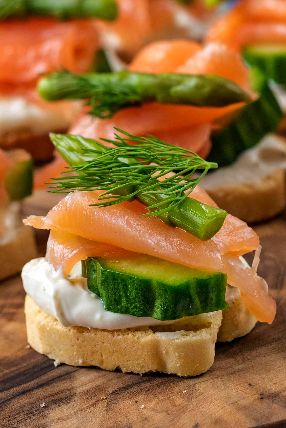 A canape made up of a crostino, some cheese, cucumber, salmon, asparagus and dill.
