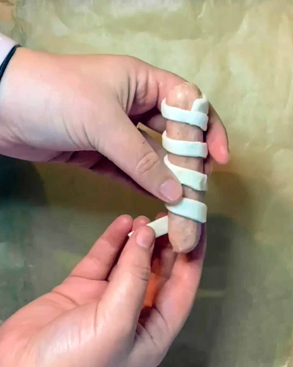 Two hands wrapping strips of pastry around a sausage.