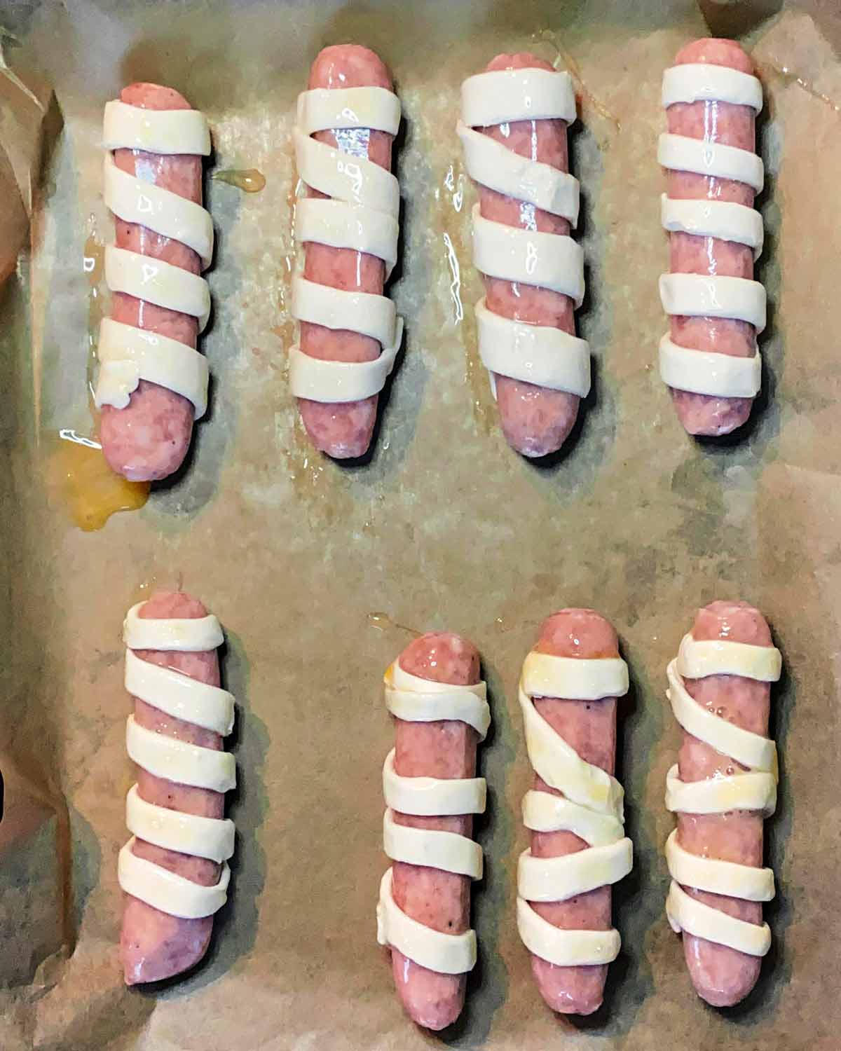 Eight sausages wrapped in strips of pastry all on a baking tray.