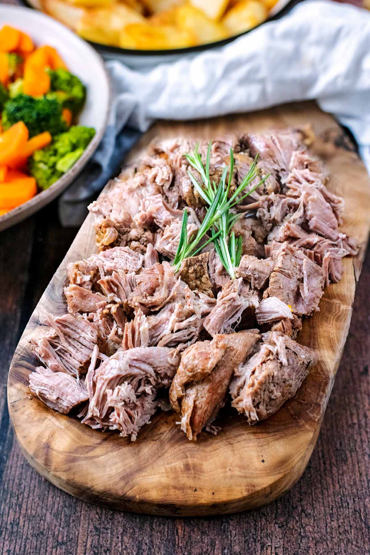 Cooked shredded lamb on a serving board in front of bowls of vegetables.