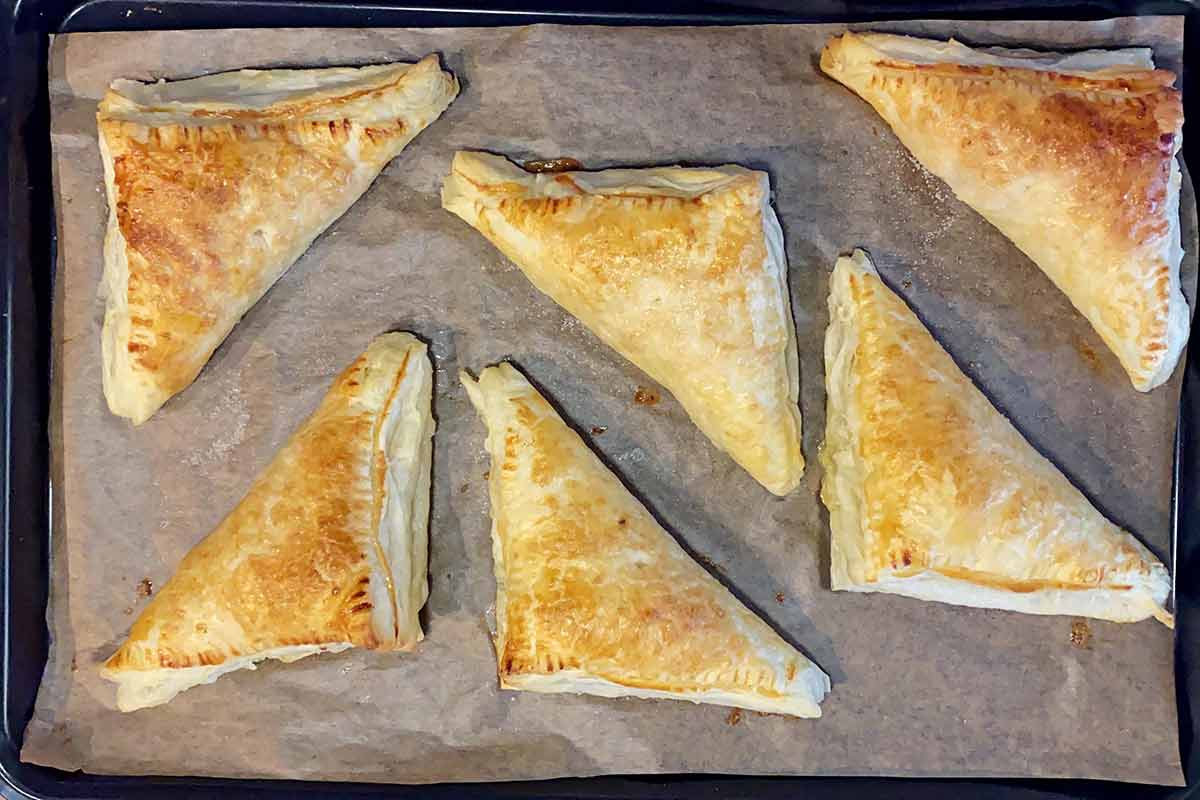 Six cooked apple turnovers on a baking tray.