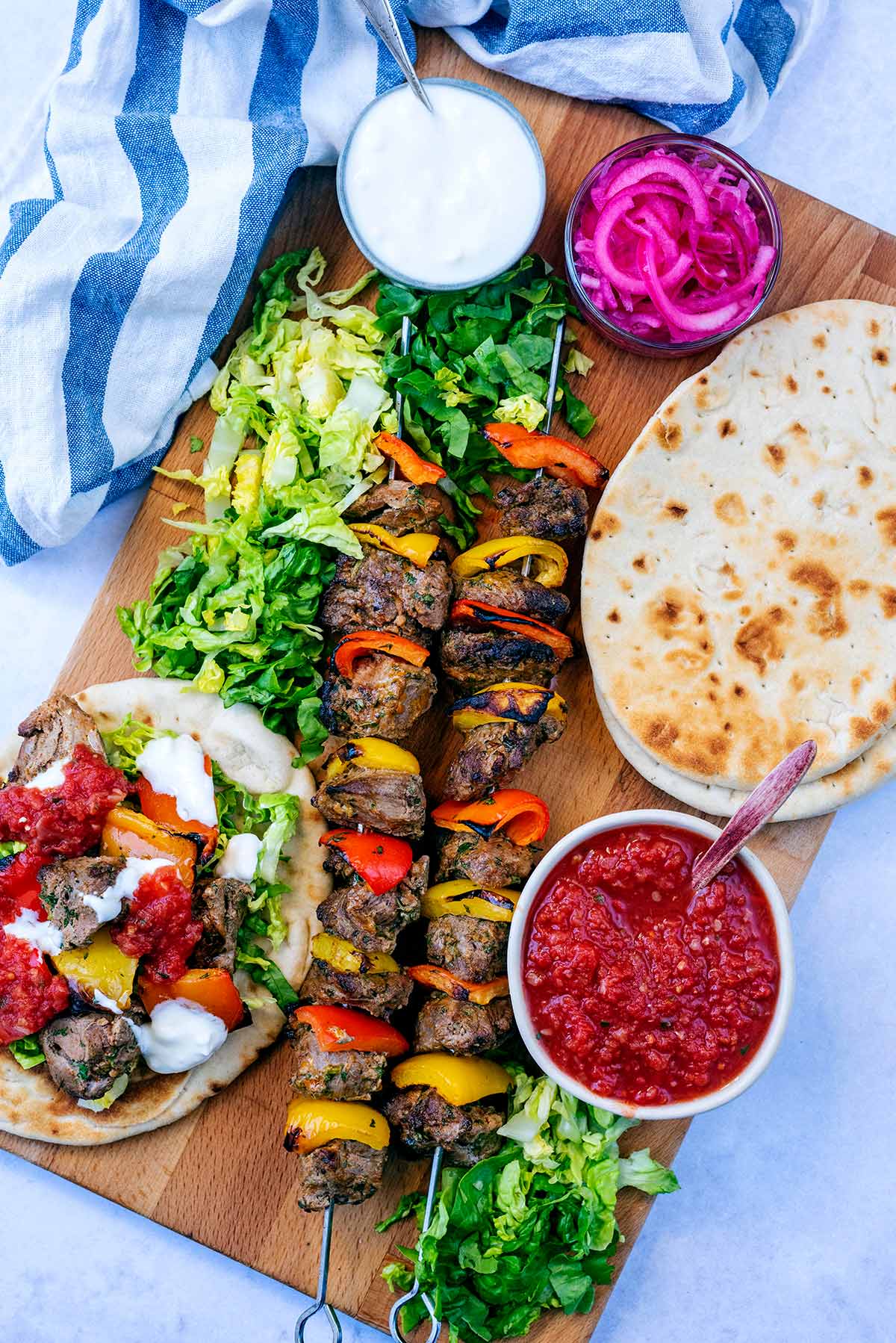 Two lamb kebab skewers on a serving board with flatbreads and sauces.