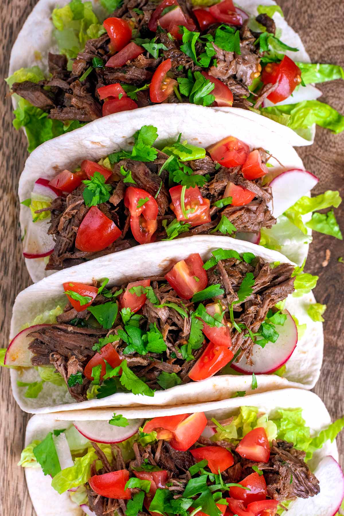 Four shredded beef barbacoa tacos topped with tomatoes and herbs.