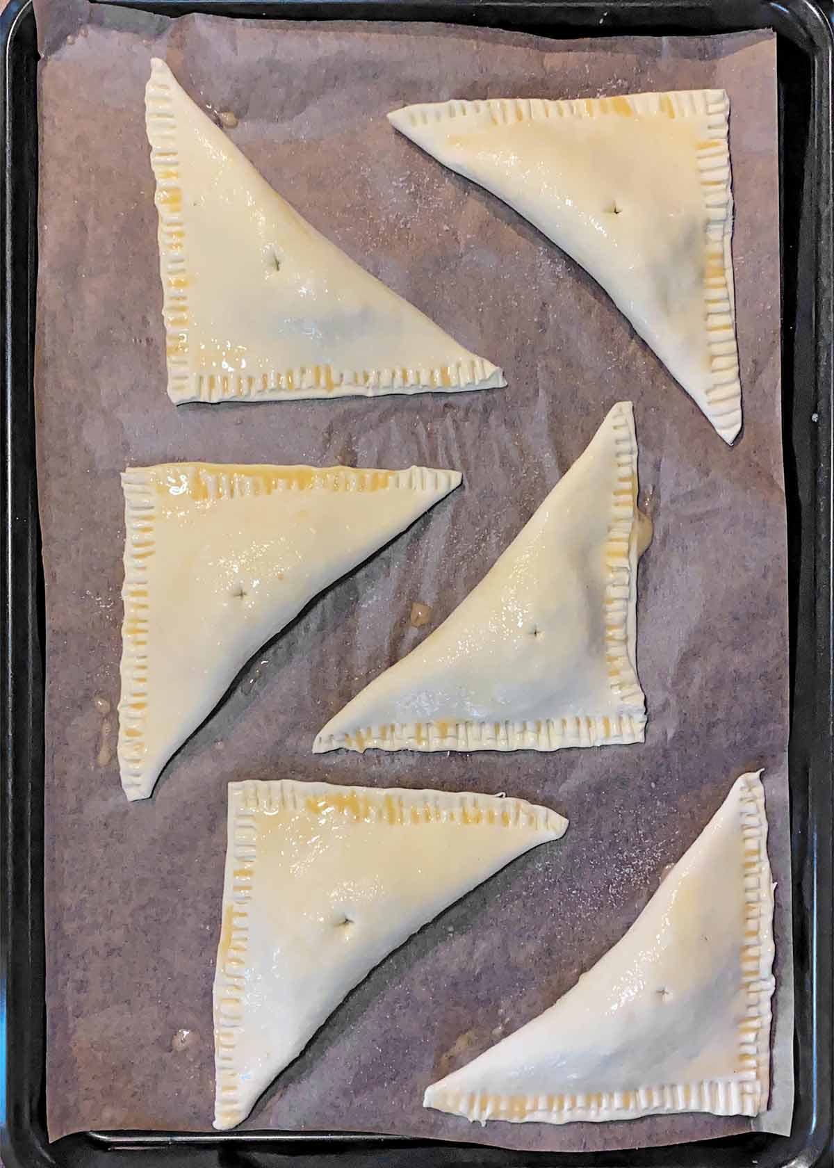 Six turnovers on a lined baking tray, each brushed with egg and sprinkled with sugar.