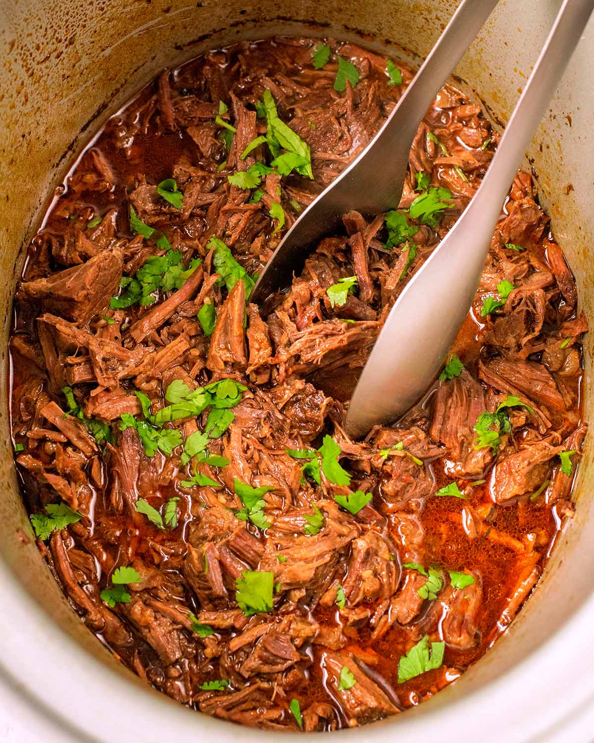 Shredded beef in a slow cooker with a set of tongs.