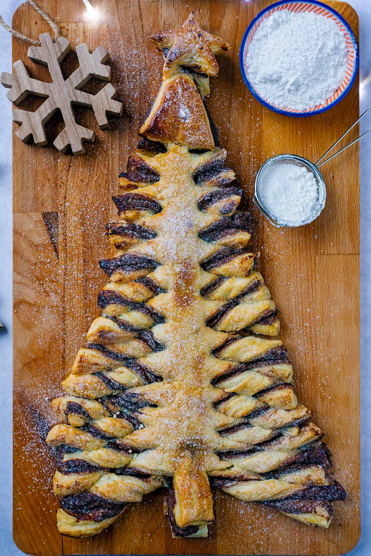A christmas tree made out of puff pastry on a board with some icing sugar and a Christmas decoration.