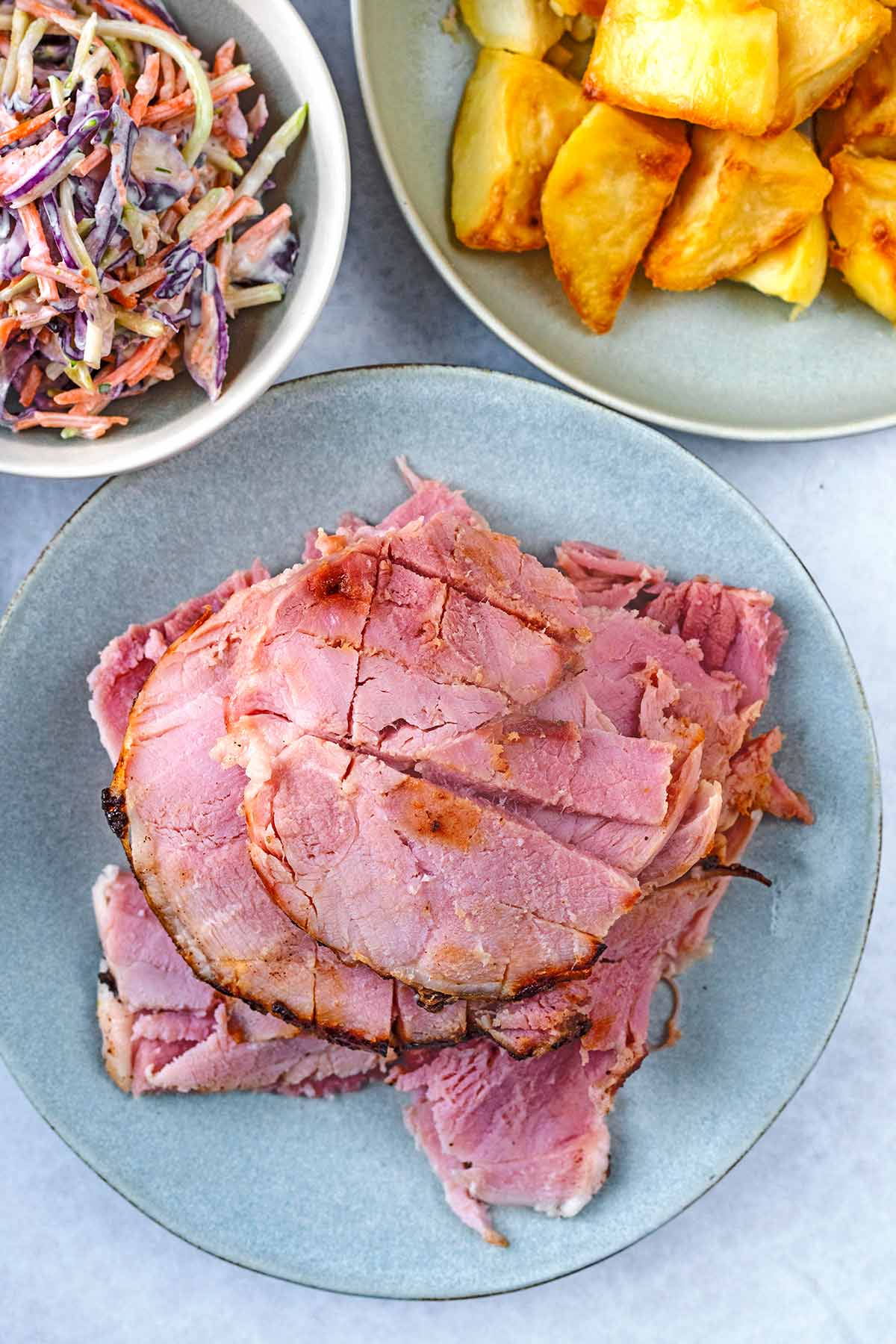 A plate of sliced ham next to bowls of roast potatoes and slaw.
