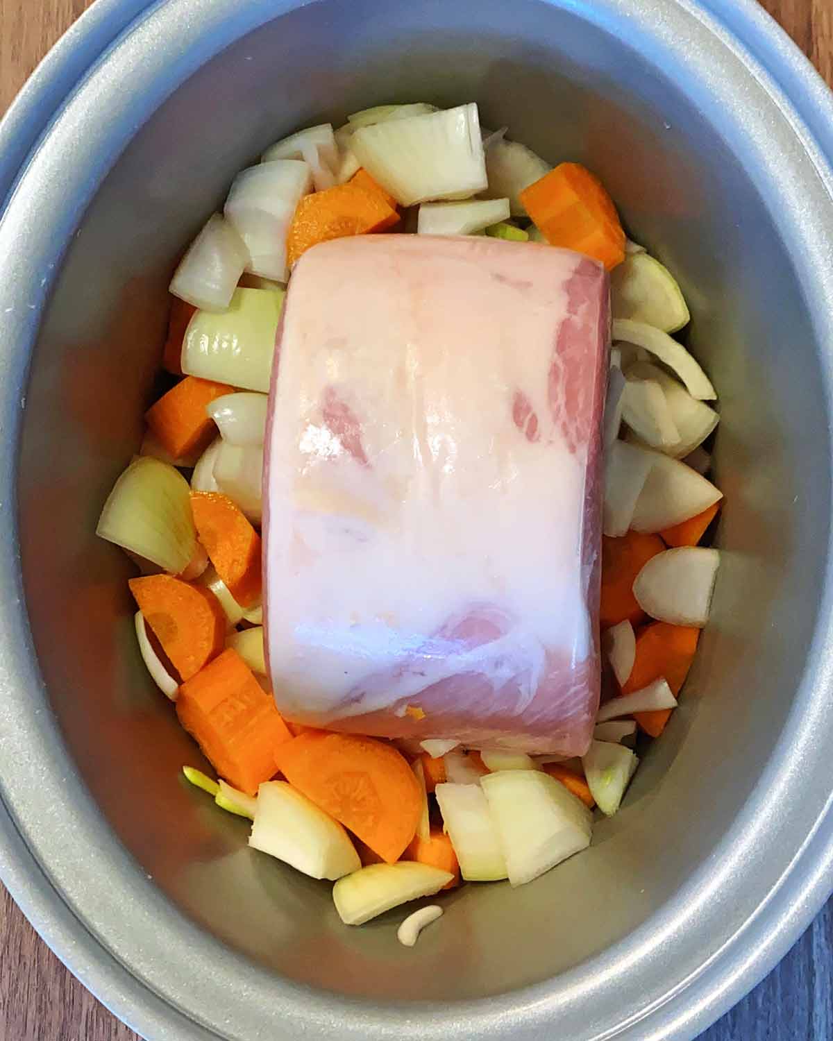 An uncooked gammon joint in a slow cooker with raw chopped vegetables.