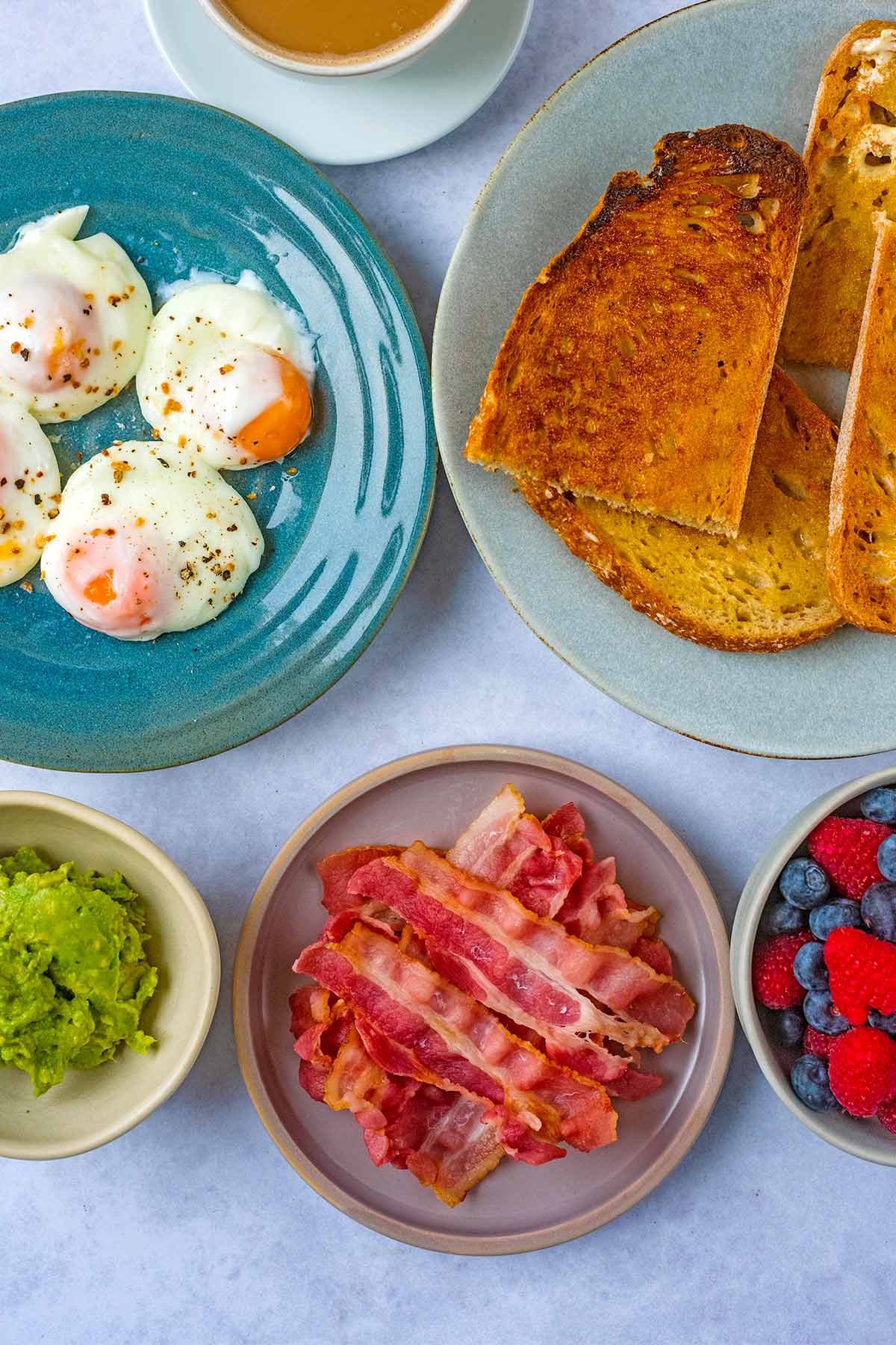 Plates of toast, eggs and bacon with bowls of smashed avocado and berries.