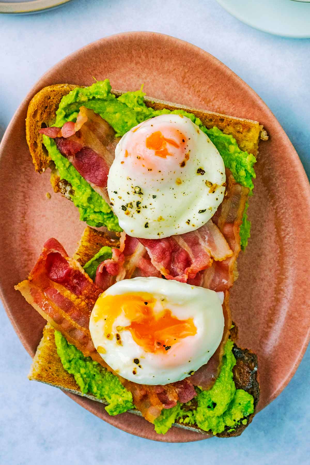 An oval plate with two slices of toast, smashed avocado, bacon and poached eggs.