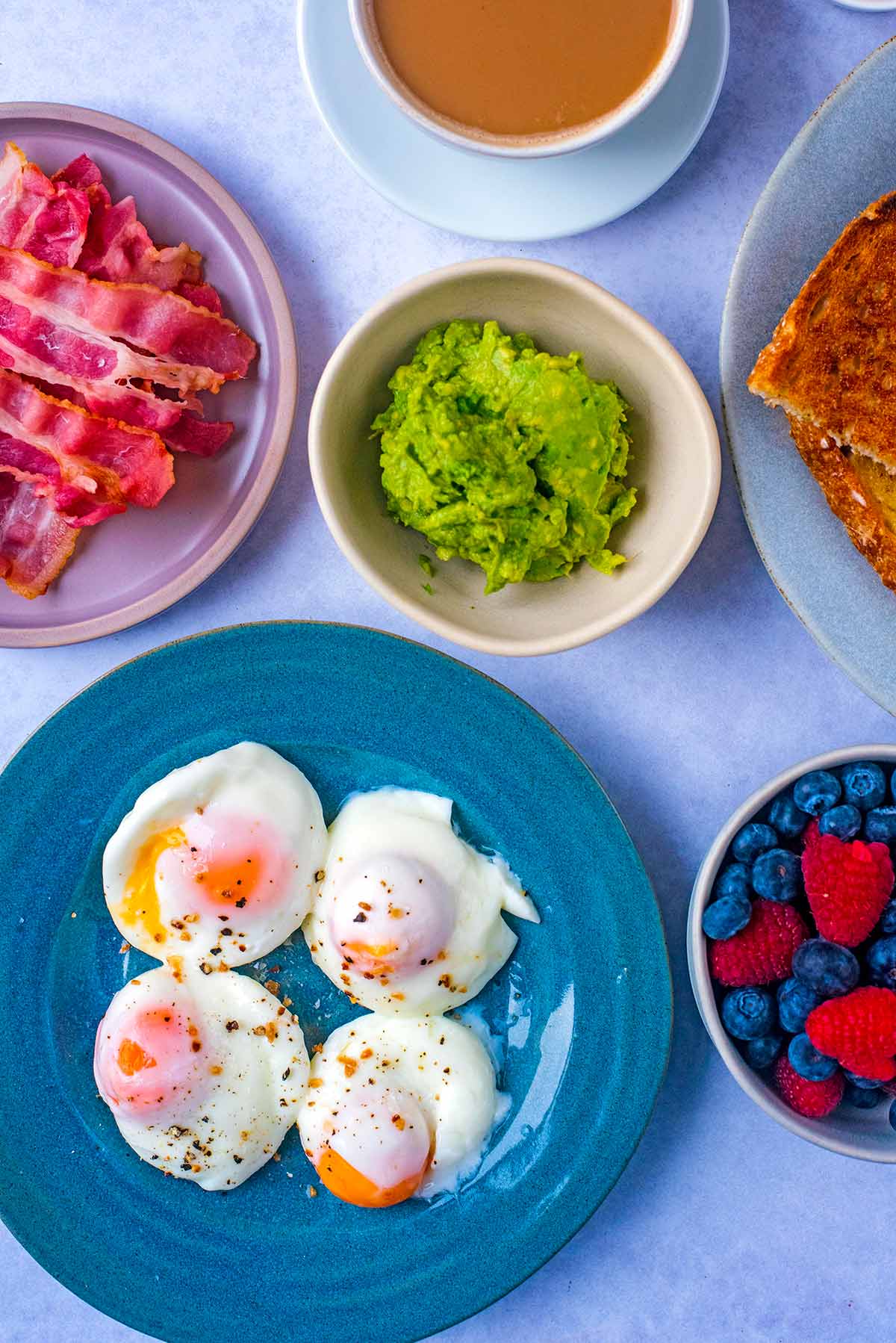 Plates of toast, poached eggs and bacon with bowls of smashed avocado and berries.