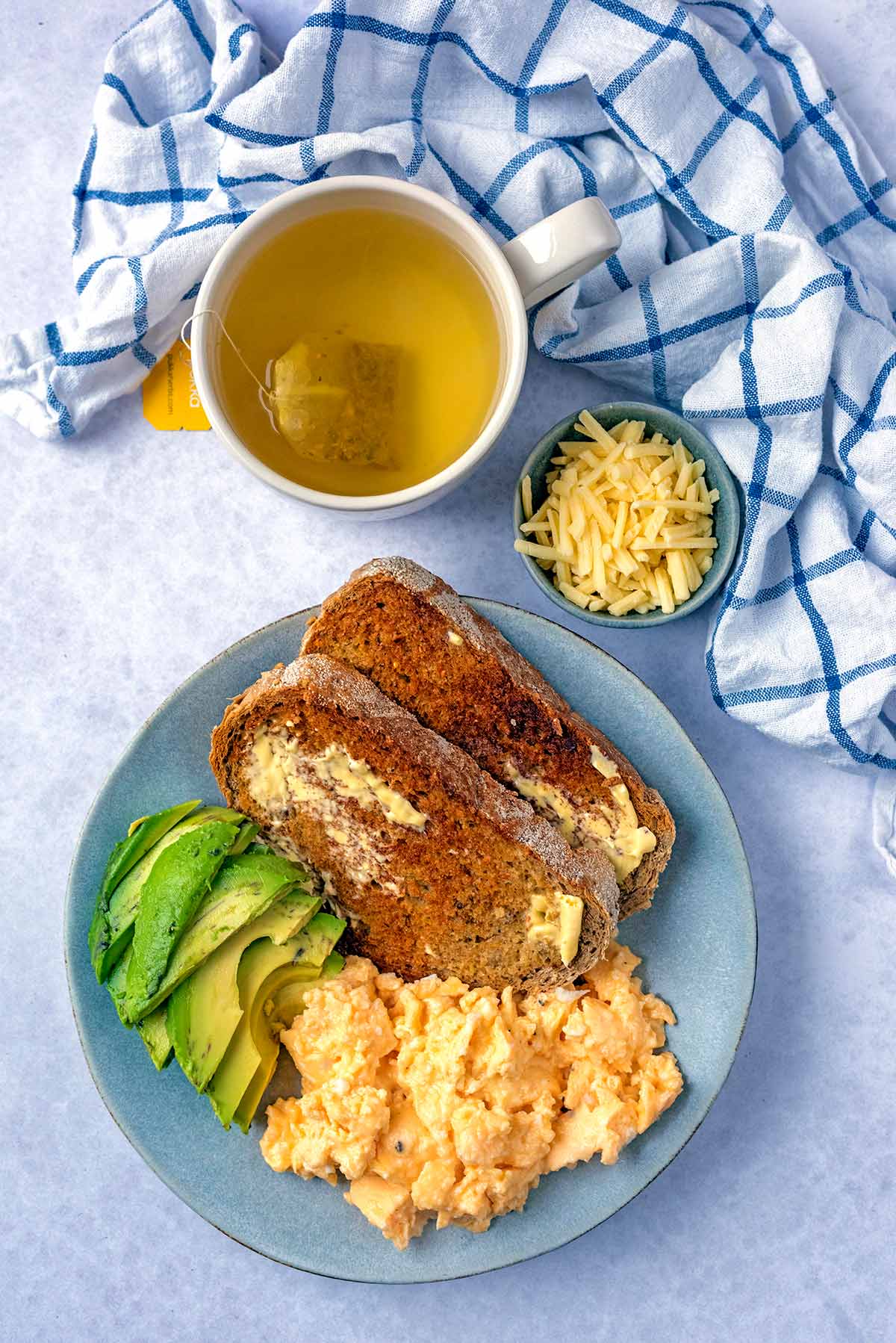 A plate of scrambled eggs, sliced avocado and toast next to a cup of tea.
