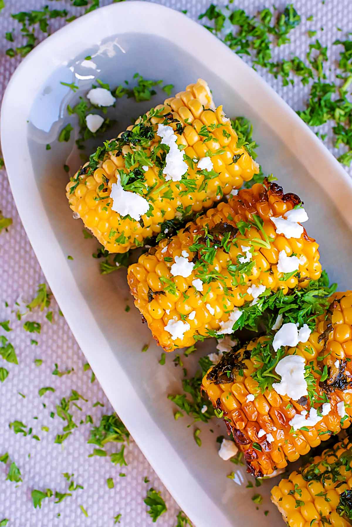 Small corn on the cobs on a long serving dish topped with chopped herbs and crumbled cheese.