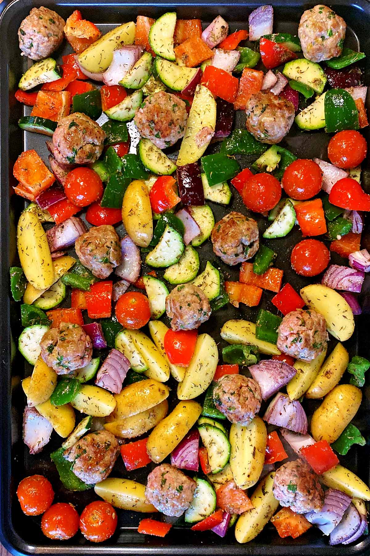 Vegetables and browned meatballs on a large black baking tray.
