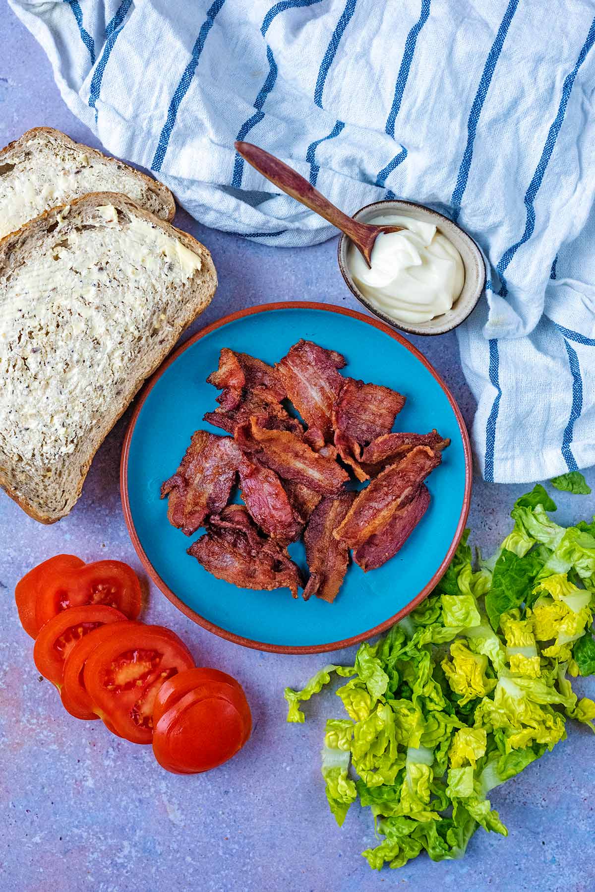 A plate of bacon next to buttered bread, lettuce and sliced tomatoes.