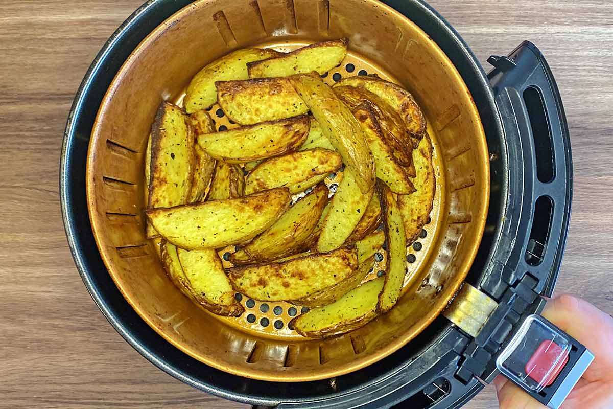 A hand holding an air fryer basket containing cooked potato wedges.