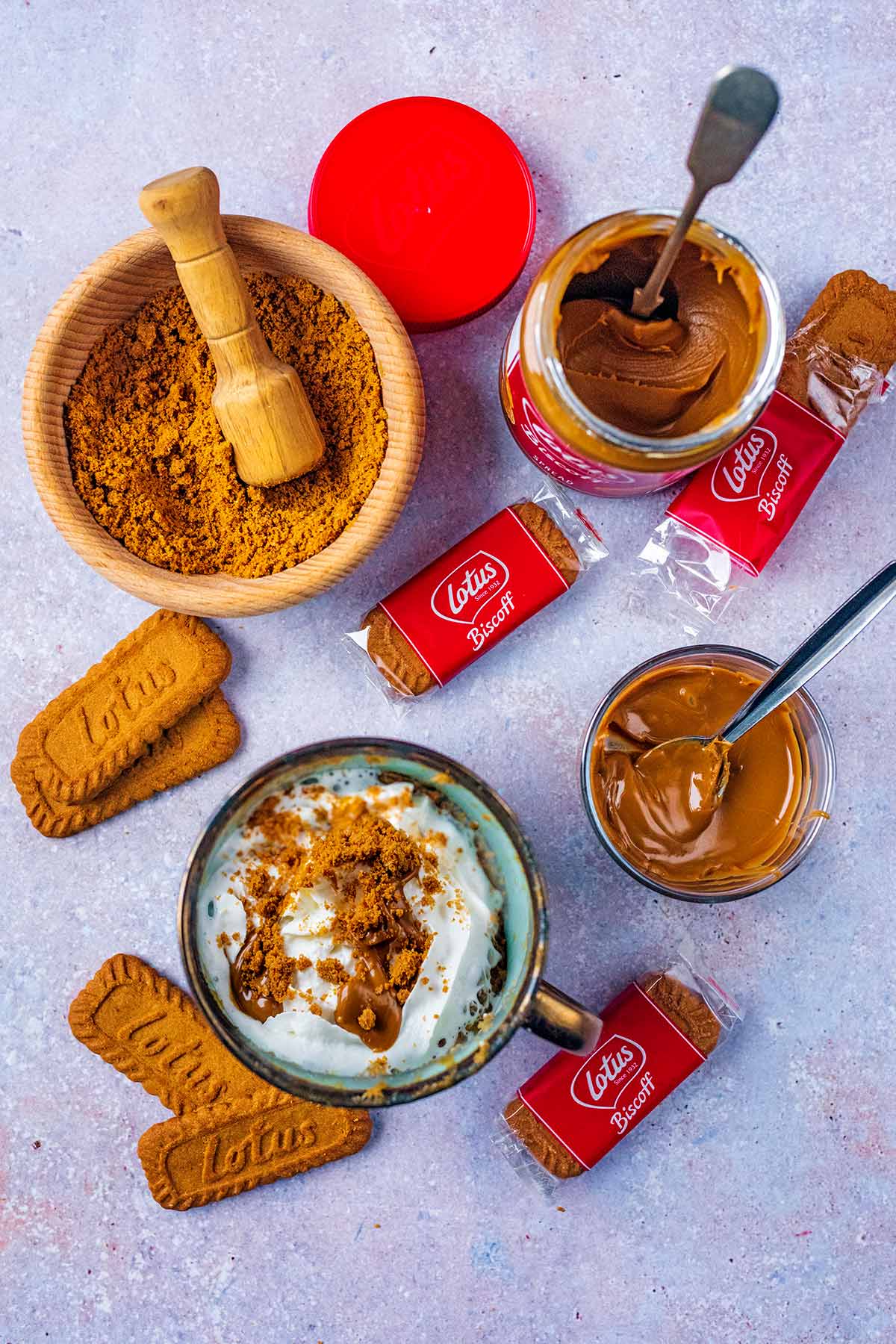 A mug cake, bowl of crushed biscuits and a jar of open Biscoff spread, all surrounded by Biscoff biscuits.