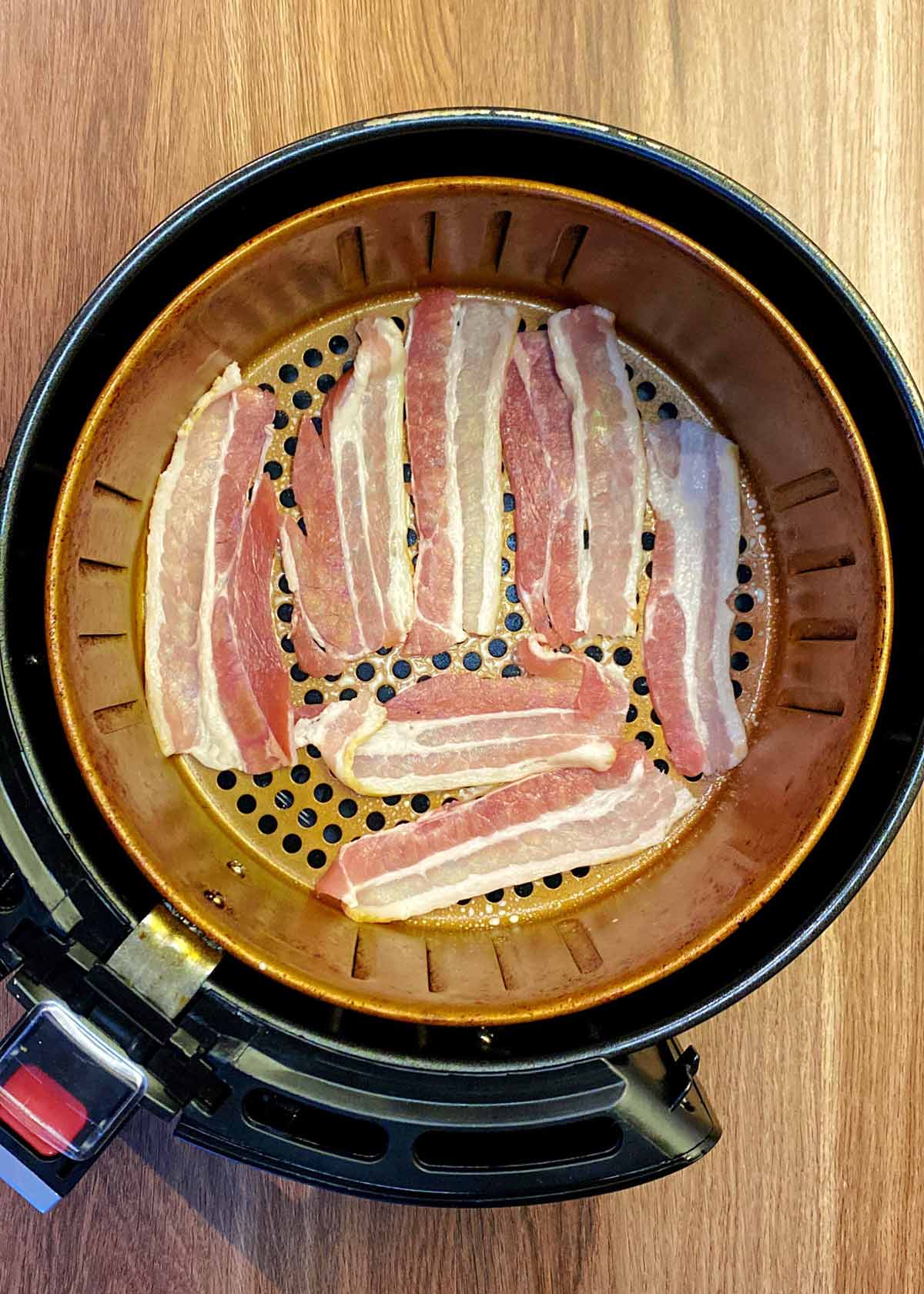 An oiled air fryer basket with rashers of uncooked bacon in it.