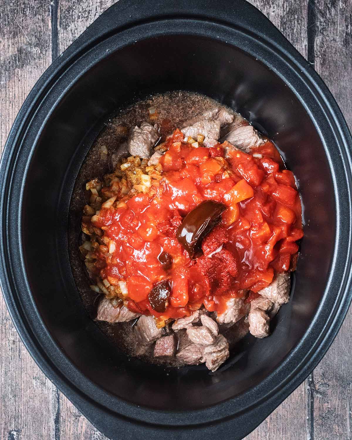 Browned beef, tomatoes, cooked onions and stock in a slow cooker bowl.