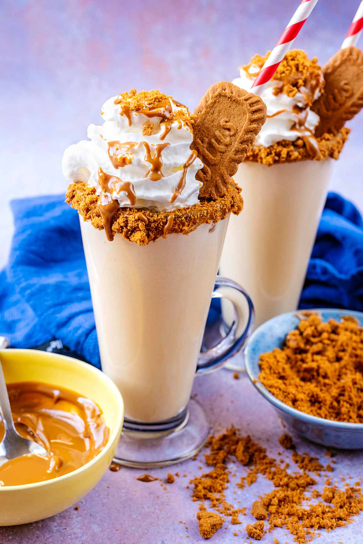 Two tall glasses of milkshake topped with cream, crushed biscuits and sauce.