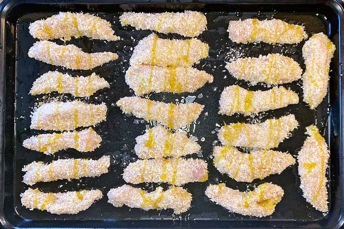 Uncooked breaded chicken strips laid onto a baking tray.