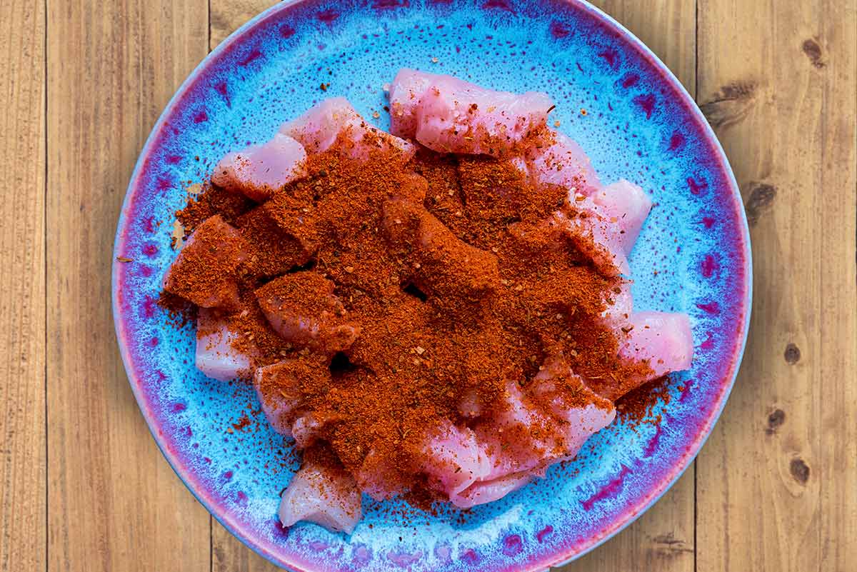 A blue plate with raw chunks of chicken breast coated in cajun seasoning.