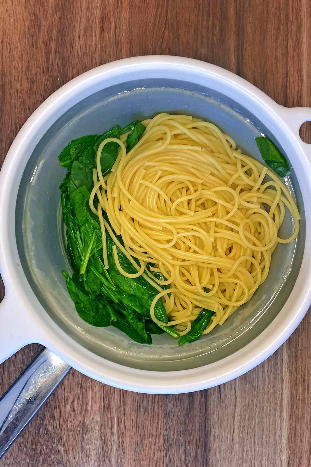 A colander containing drained spaghetti and wilted spinach leaves. 
