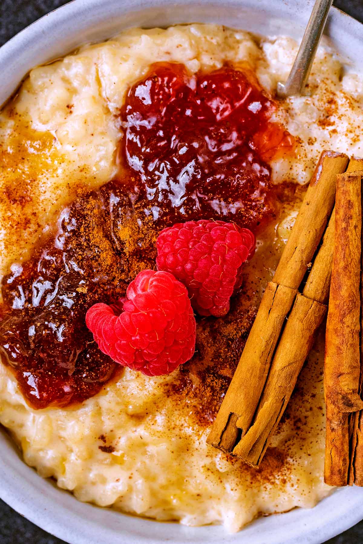 A dollop of jam, two raspberries and some cinnamon sticks on top of some rice pudding.