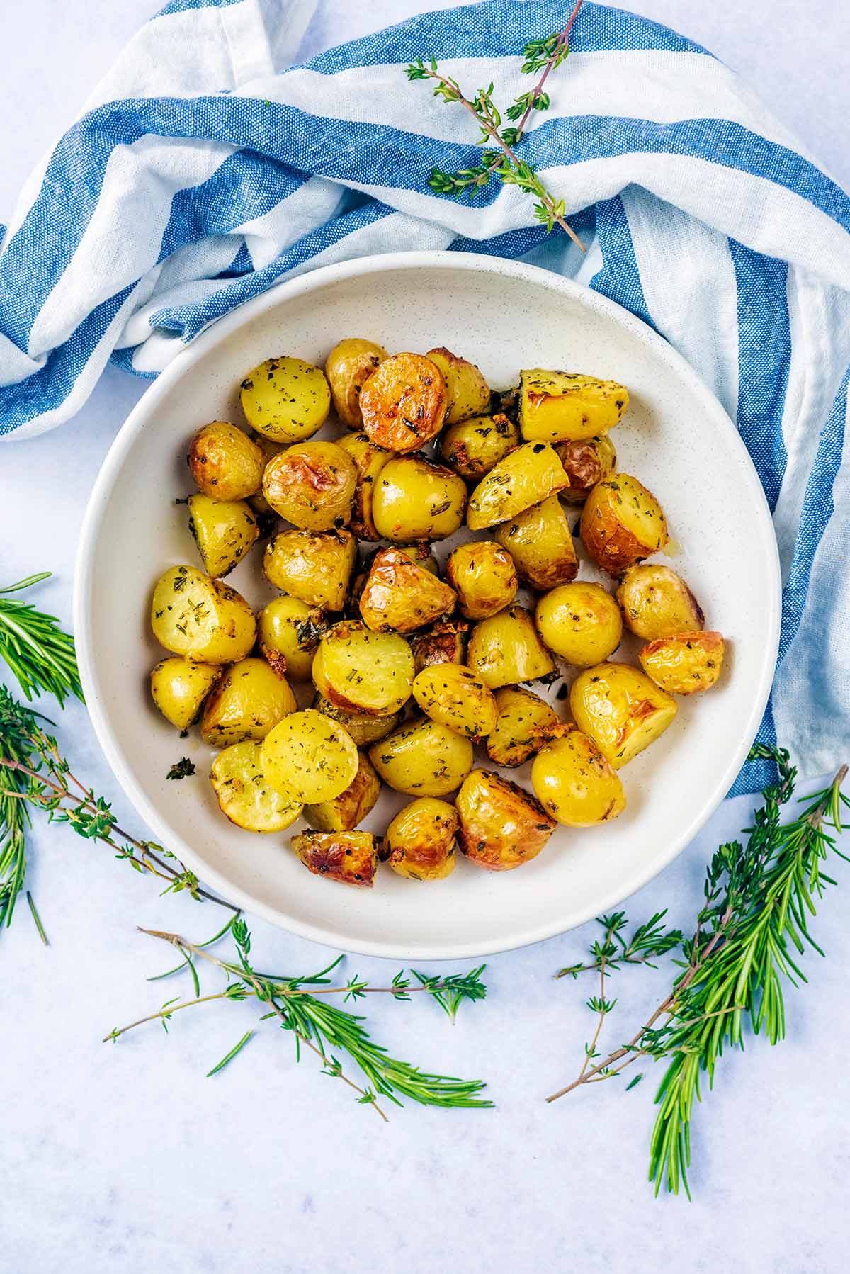 A bowl of roast baby potatoes next to sprigs of rosemary and thyme.