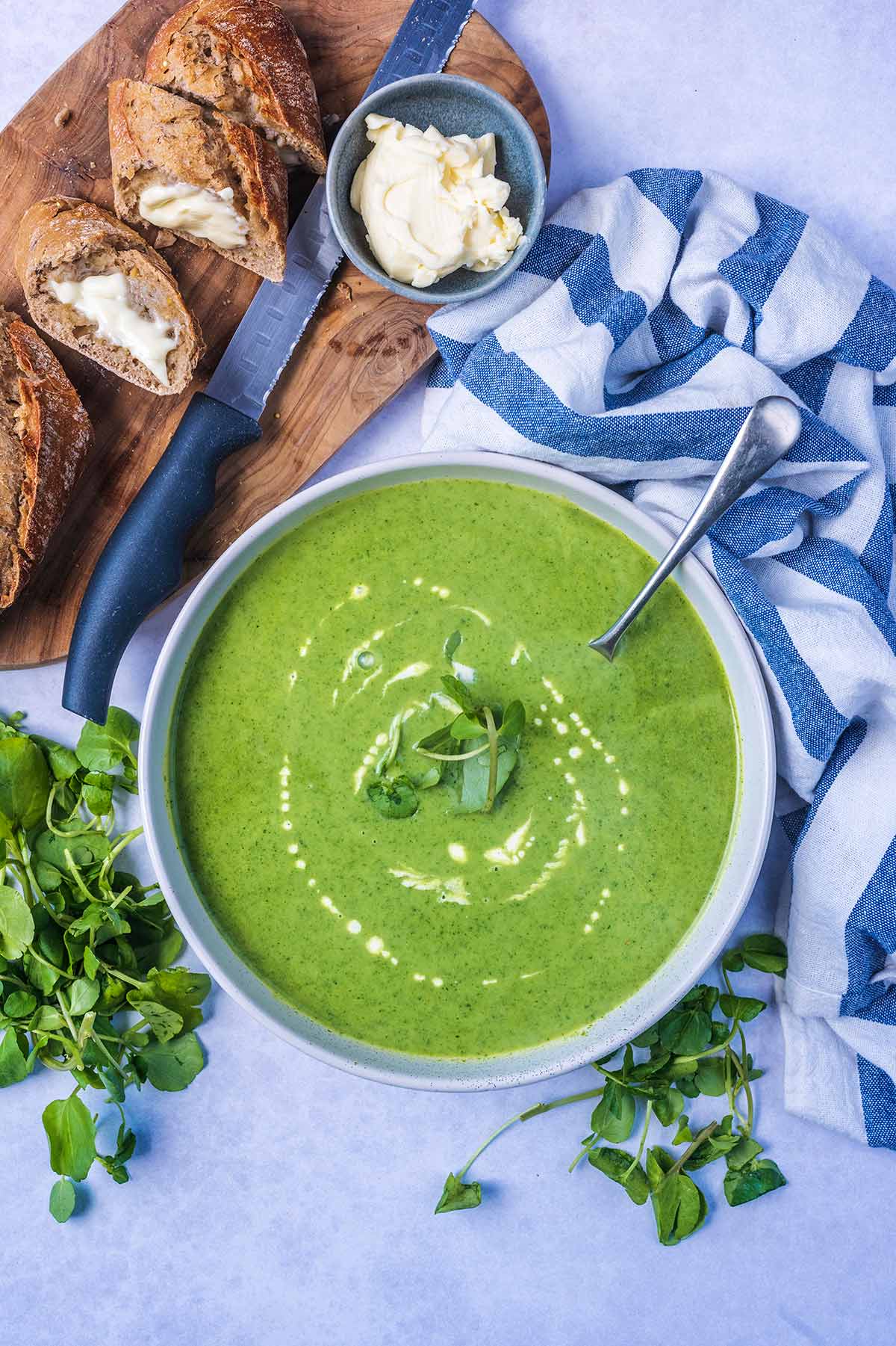 A bowl of green soup next to a towel and some sliced bread.