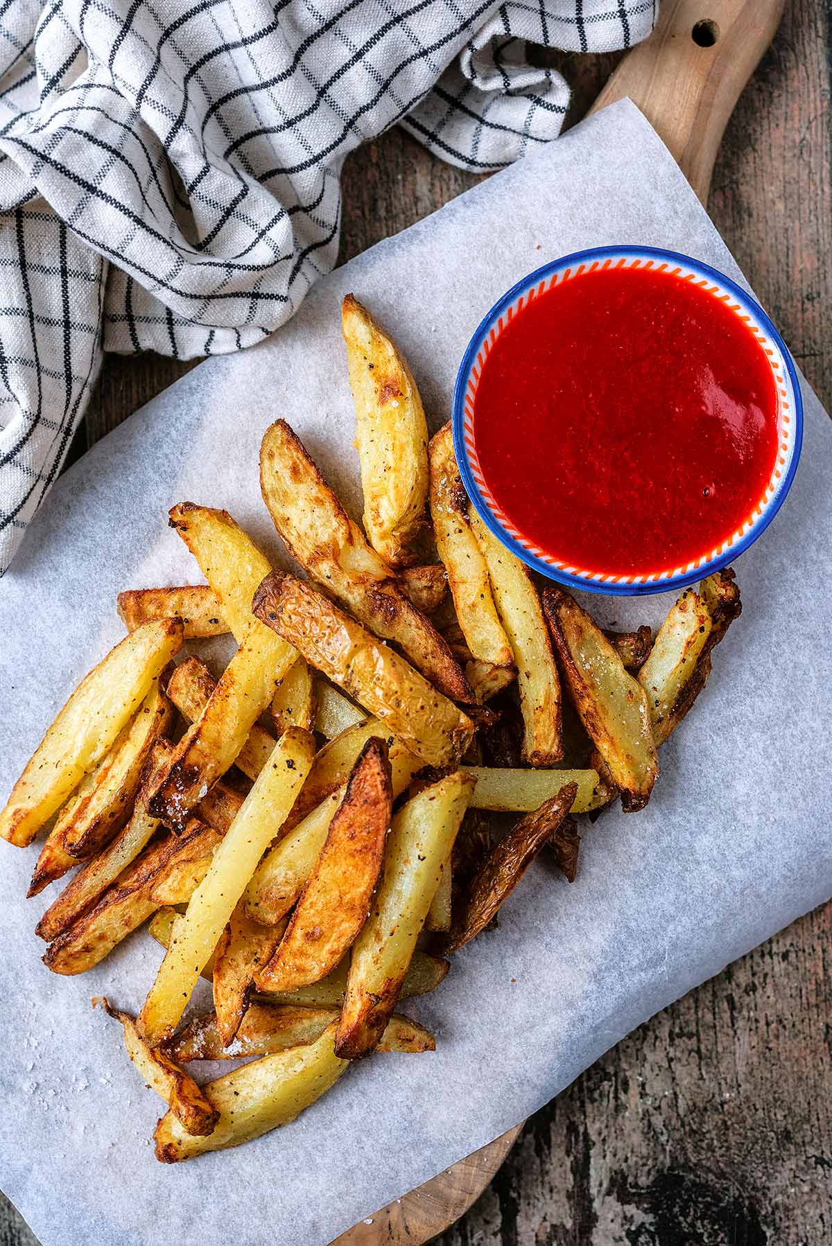 Potato chips on a wooden serving board with a small pot of ketchup.