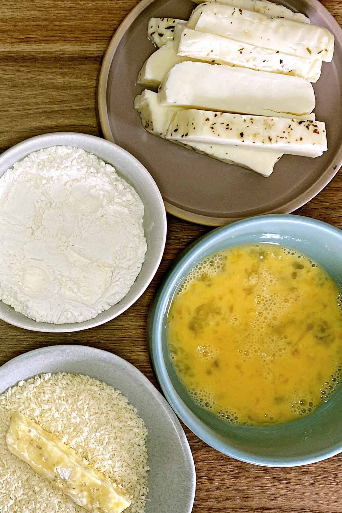 Strips of halloumi next to bowls of flour, breadcrumbs and whisked egg.