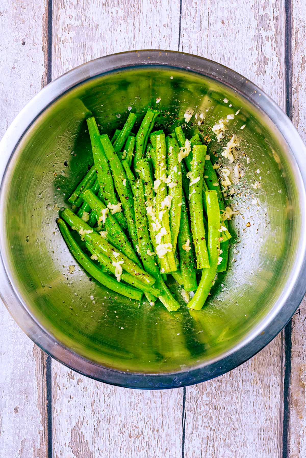 A mixing bowl with green beans, oil and crushed garlic.