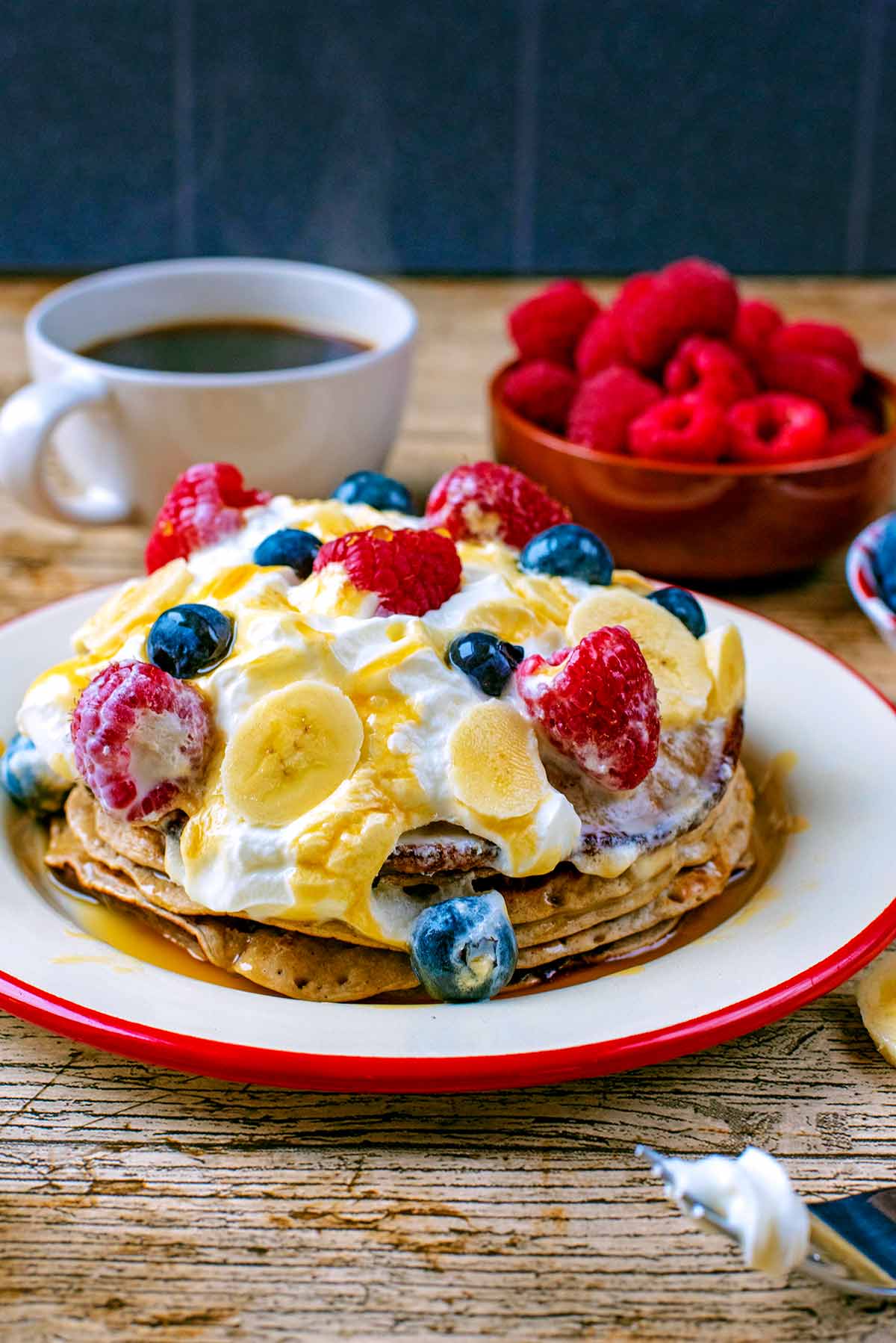 Pancakes topped with cream on a plate. A cup of coffee and raspberries in the background.