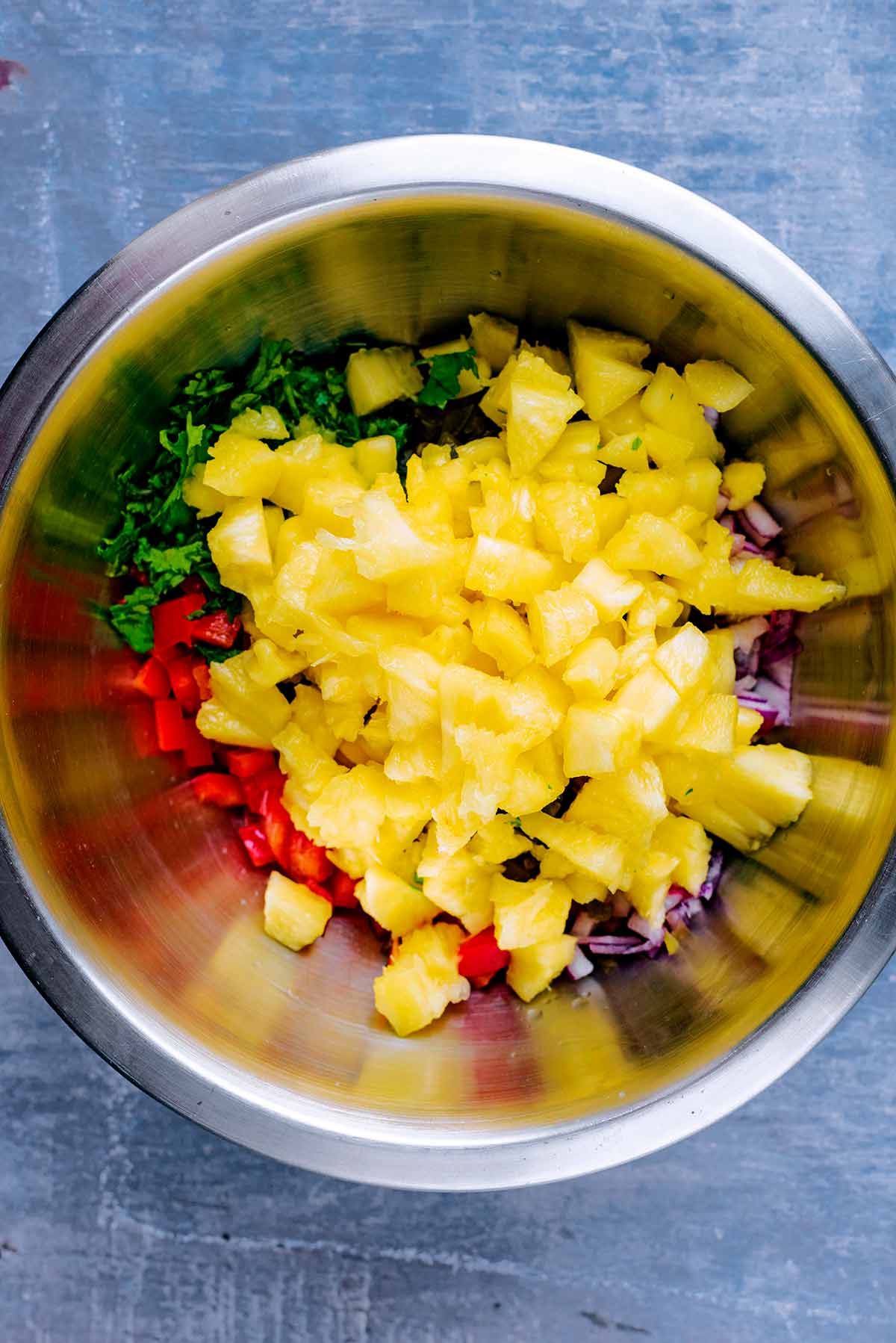 A mixing bowl containing pineapple chunks, chopped bell pepper and red.onion and chopped herbs.