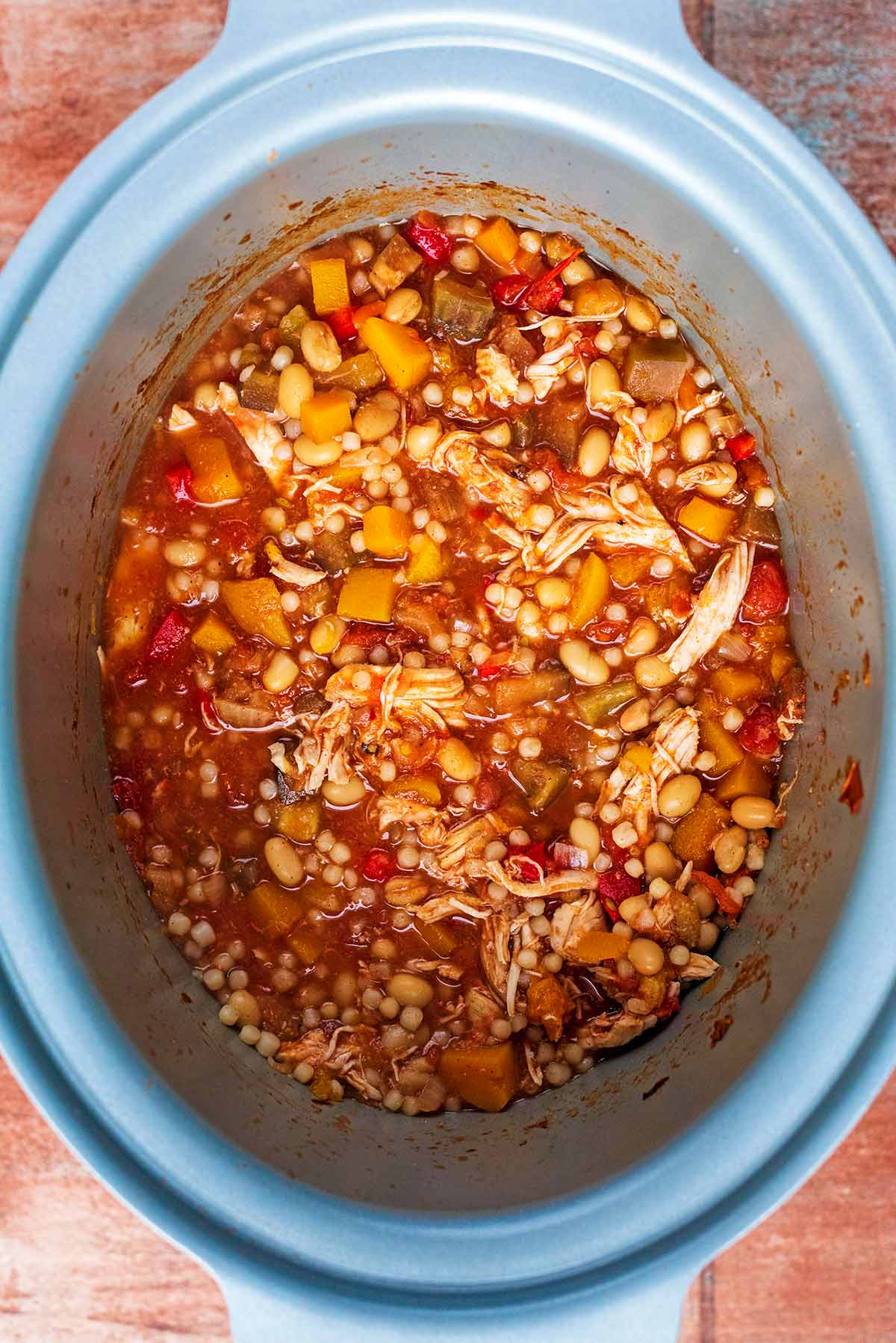 A slow cooker bowl containing shredded chicken, chopped vegetables, beans and couscous.