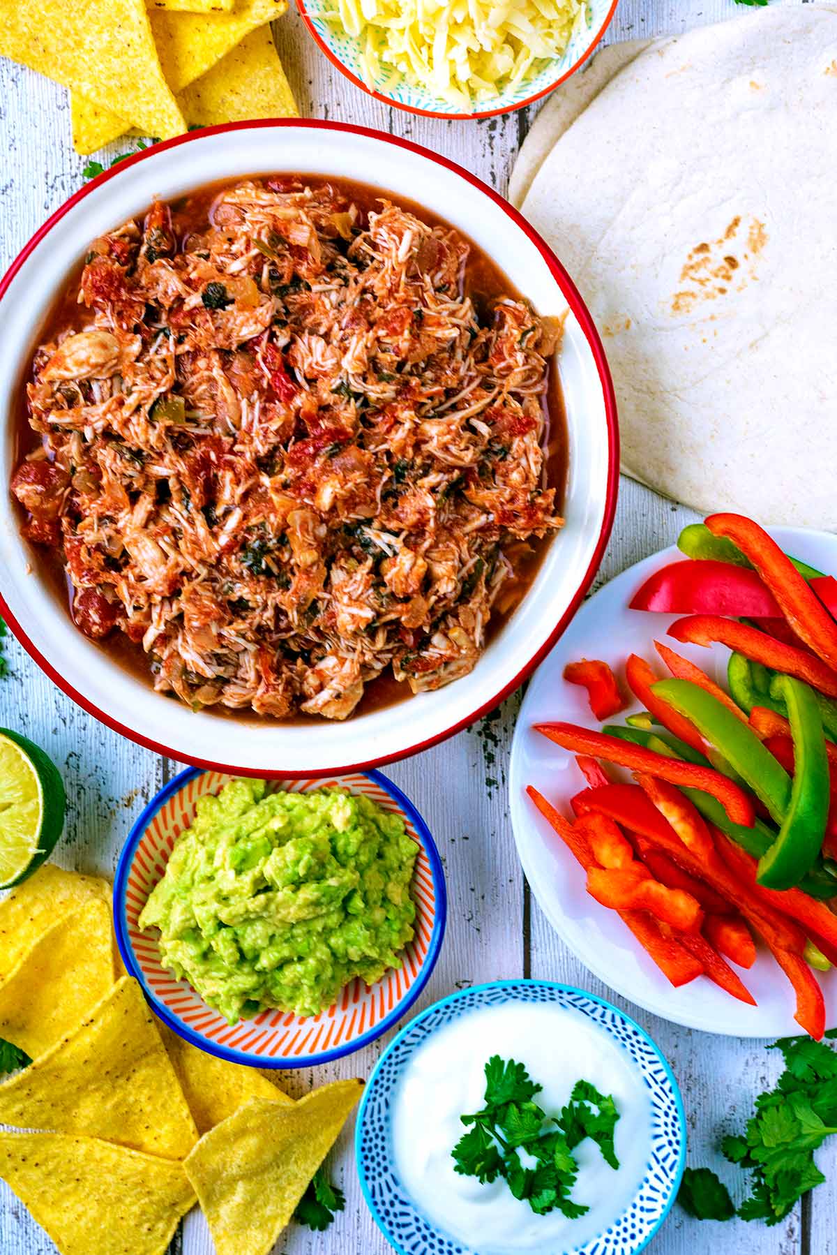 Salsa chicken in a dish next to some guacamole, flour tortillas, sliced peppers, sour cream and nachos.