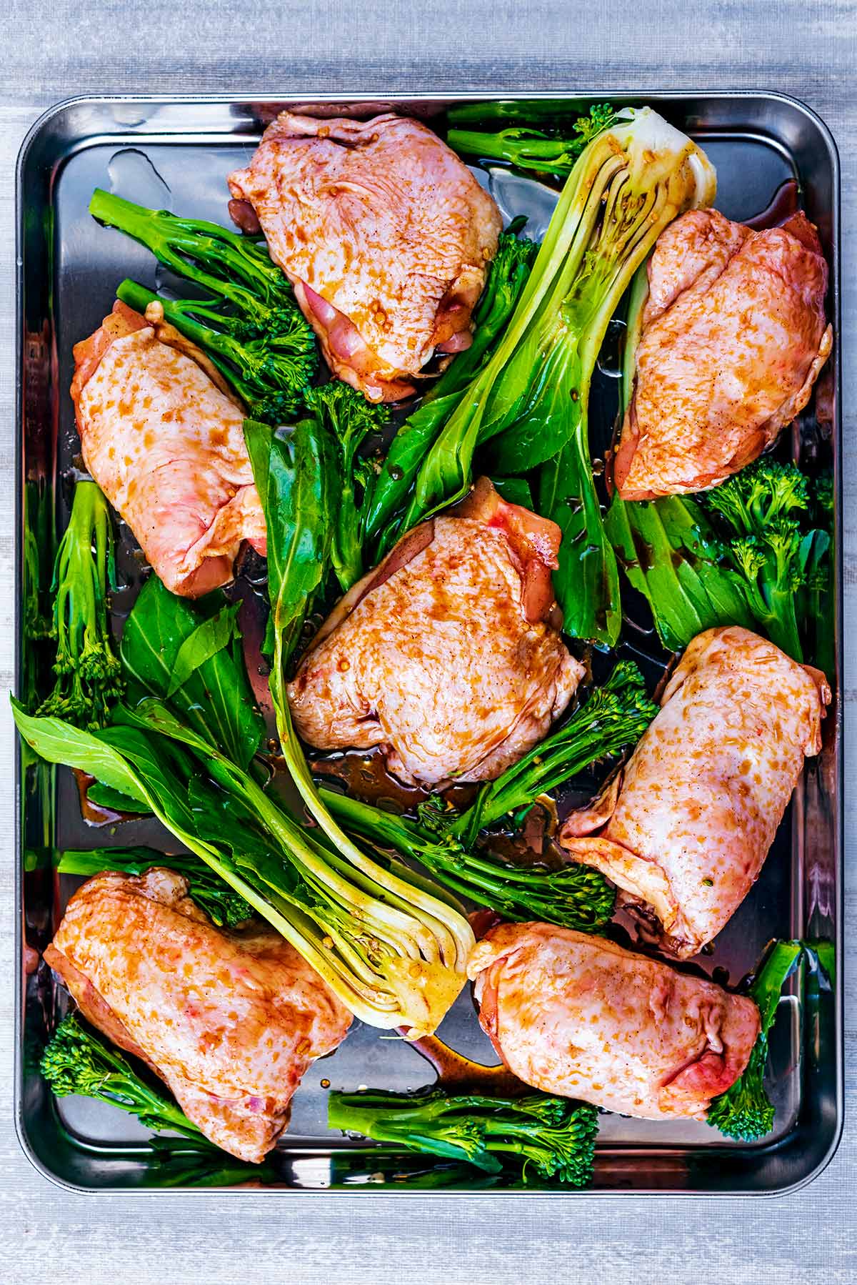 A large baking tray with seven raw marinated chicken thighs and halves of pak choi.