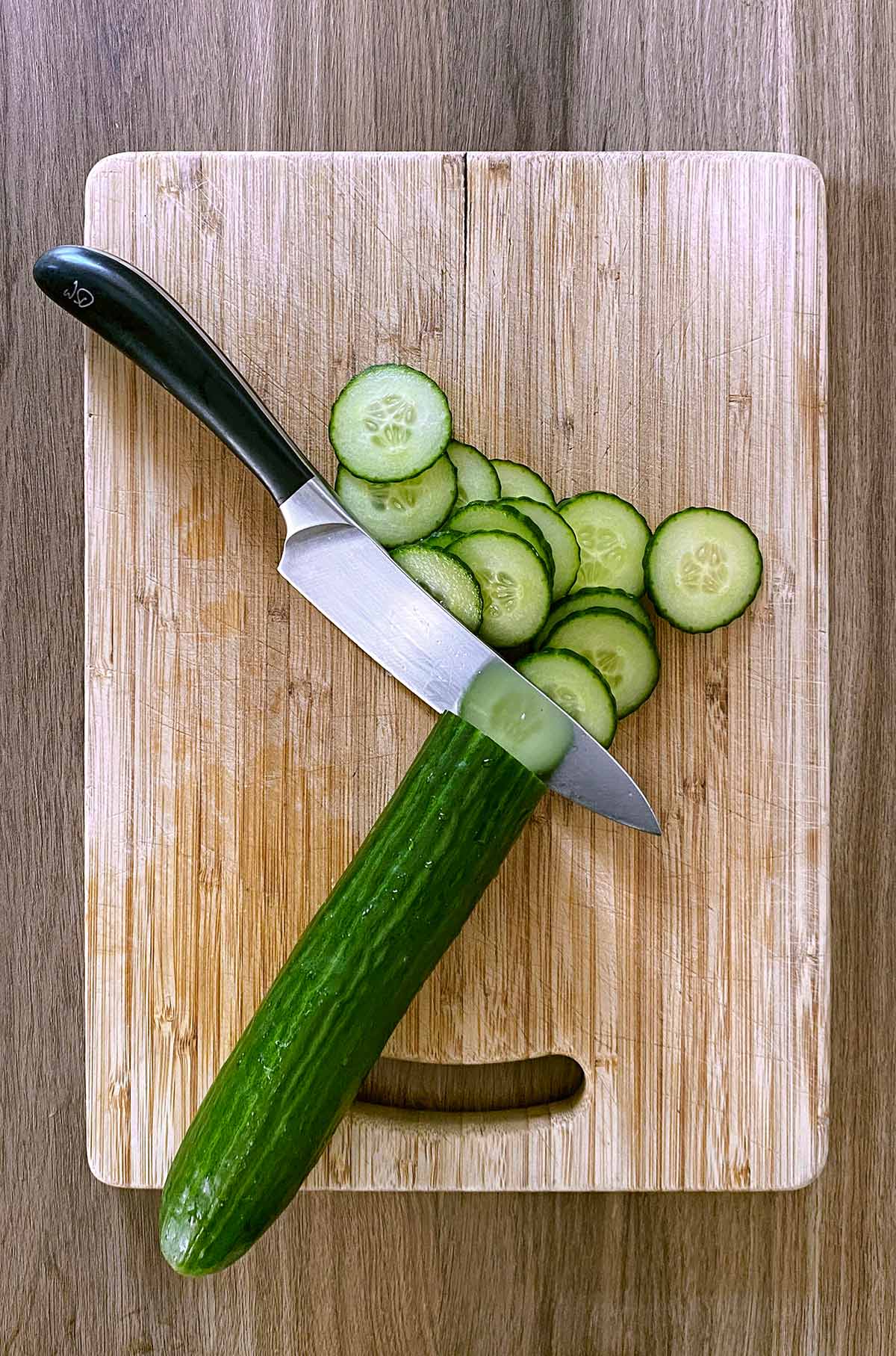 A chopping board and knife with a cucumber cut into slices.