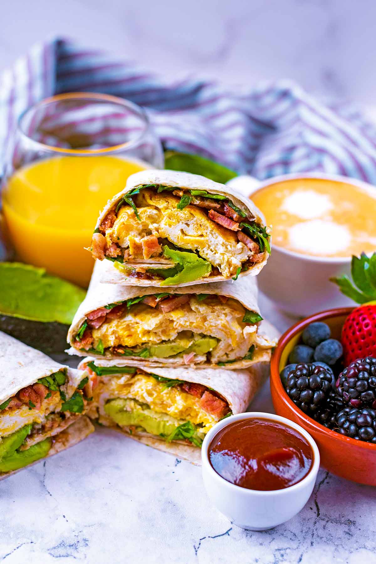 A pile of breakfast wraps next to fruit, orange juice, coffee and barbecue sauce.