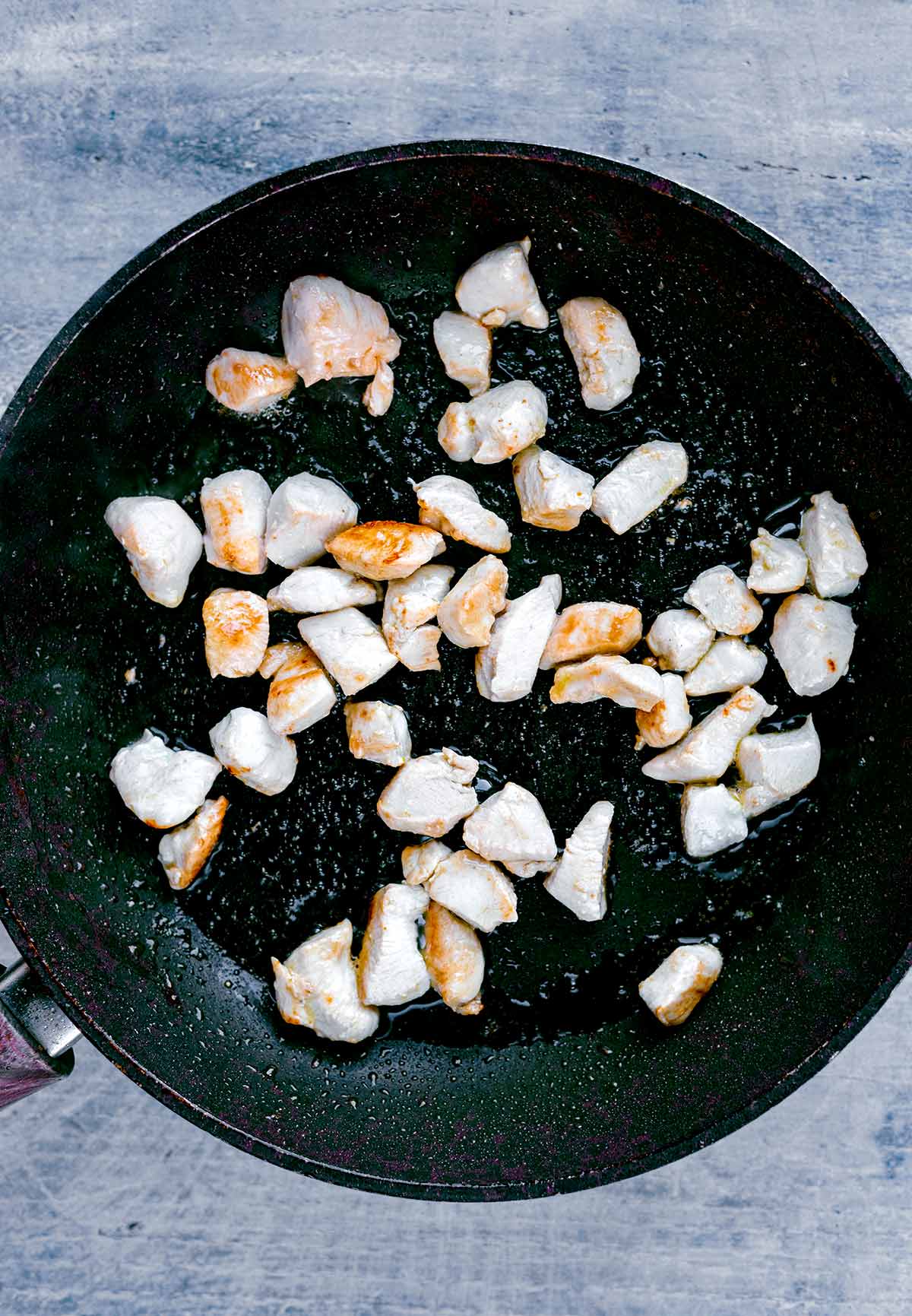 A frying pan with chunks of chicken breast cooking in it.