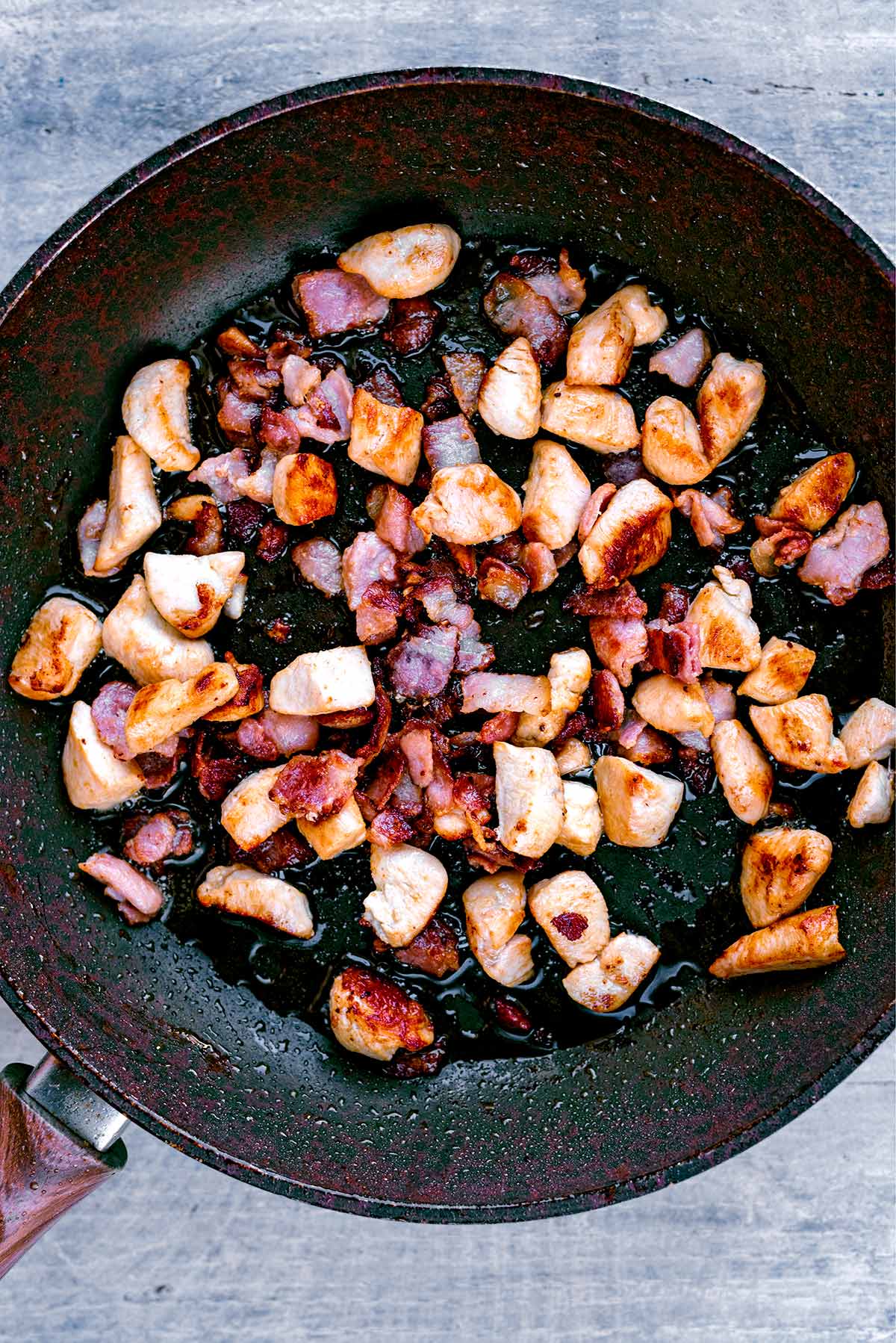 Chunks of chicken and chopped bacon cooking in a frying pan.