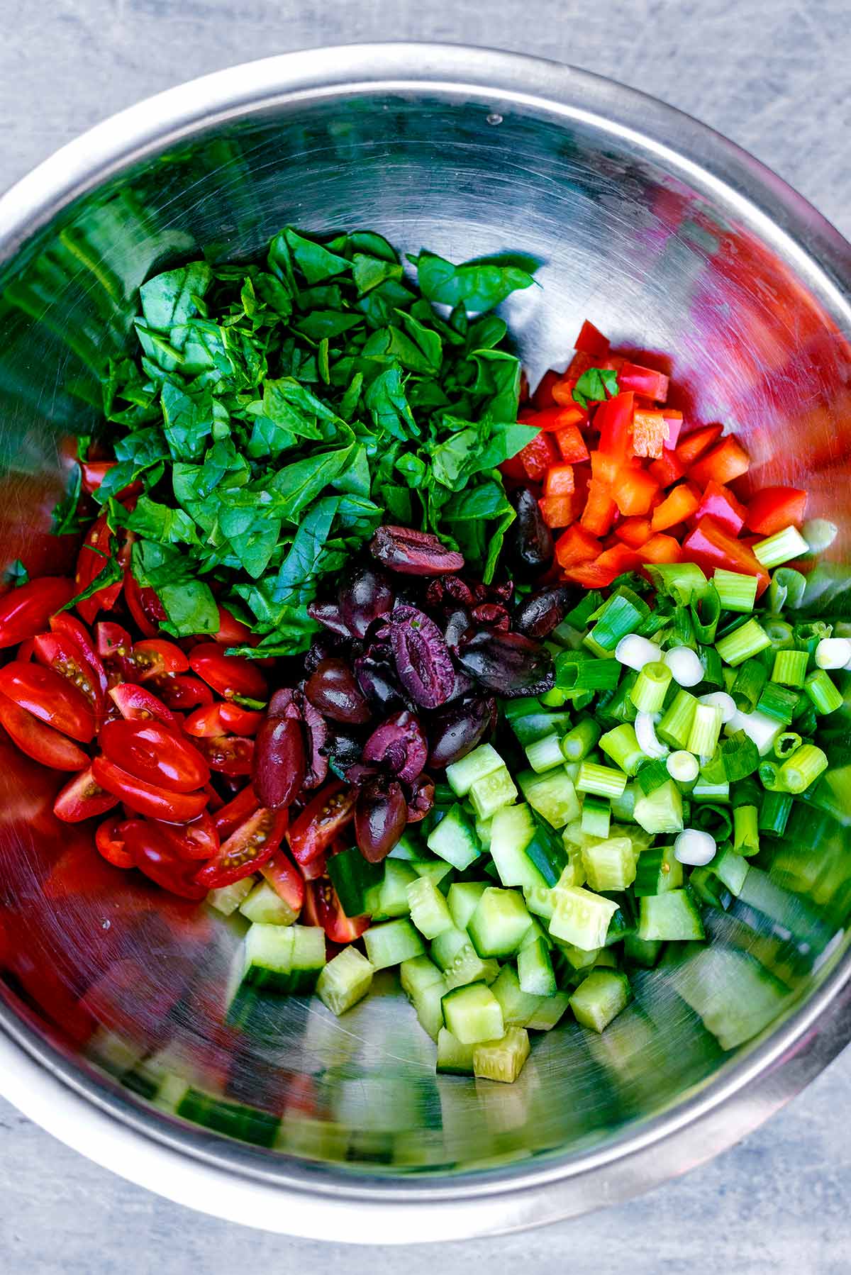 A stainless steel mixing bowl containing tomatoes, spinach, red pepper, onions, cucumber and olives.
